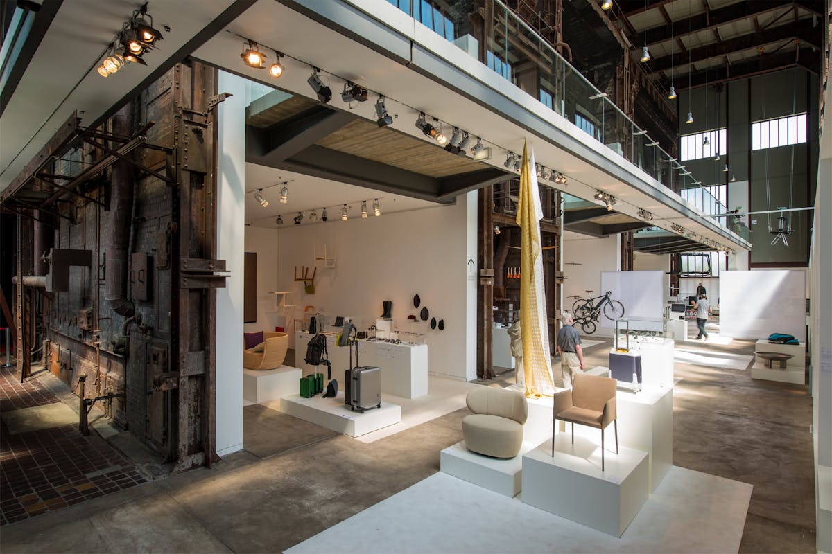 Ein Blick in das Red Dot Design Museum auf dem UNESCO-Welterbe Zollverein. Die Ausstellung verbindet moderne Design-Objekte mit der historischen Industriearchitektur der ehemaligen Zeche. Links im Bild sind rostige Stahlwände, Rohre und Metallstrukturen der alten Industrieanlage sichtbar, während die rechte Seite von einer offenen, hellen Ausstellungsfläche dominiert wird. Auf weißen Podesten werden preisgekrönte Designstücke präsentiert, darunter moderne Möbel, Reisekoffer, Rucksäcke, Brillen und Fahrräder. Tageslicht strömt durch hohe Fenster und hebt den Kontrast zwischen industriellem Erbe und zeitgenössischem Design hervor. Besucher bewegen sich durch die Ausstellung und erkunden die innovativen Objekte.