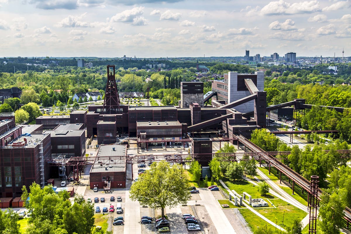 Eine beeindruckende Luftaufnahme des UNESCO-Welterbes Zollverein in Essen. Im Zentrum des Bildes steht der markante Förderturm der ehemaligen Zeche Zollverein, umgeben von den symmetrisch angeordneten, dunklen Backsteingebäuden des Industriekomplexes. Das Gelände ist durchzogen von alten Rohrleitungen und Förderbändern, die einst für den Kohletransport genutzt wurden. Im Vordergrund befinden sich Grünflächen mit Bäumen und Wiesen, die Teil des Zollverein Parks sind – einer modernen, naturnahen Umgestaltung des ehemaligen Bergbaugeländes. Parkplätze mit geparkten Autos sowie Wege für Besucher und Anwohner sind sichtbar. Im Hintergrund erstreckt sich die Skyline von Essen mit hohen Gebäuden und Kirchtürmen, eingerahmt von dicht bewachsenen, grünen Flächen, die einen starken Kontrast zur industriellen Architektur der Zeche bilden. Die helle Tagesbeleuchtung und der blaue Himmel mit weißen Wolken verleihen dem Bild eine offene, einladende Atmosphäre, die die erfolgreiche Transformation von Zollverein von einer ehemaligen Zeche zu einem Kultur- und Erholungsort unterstreicht.