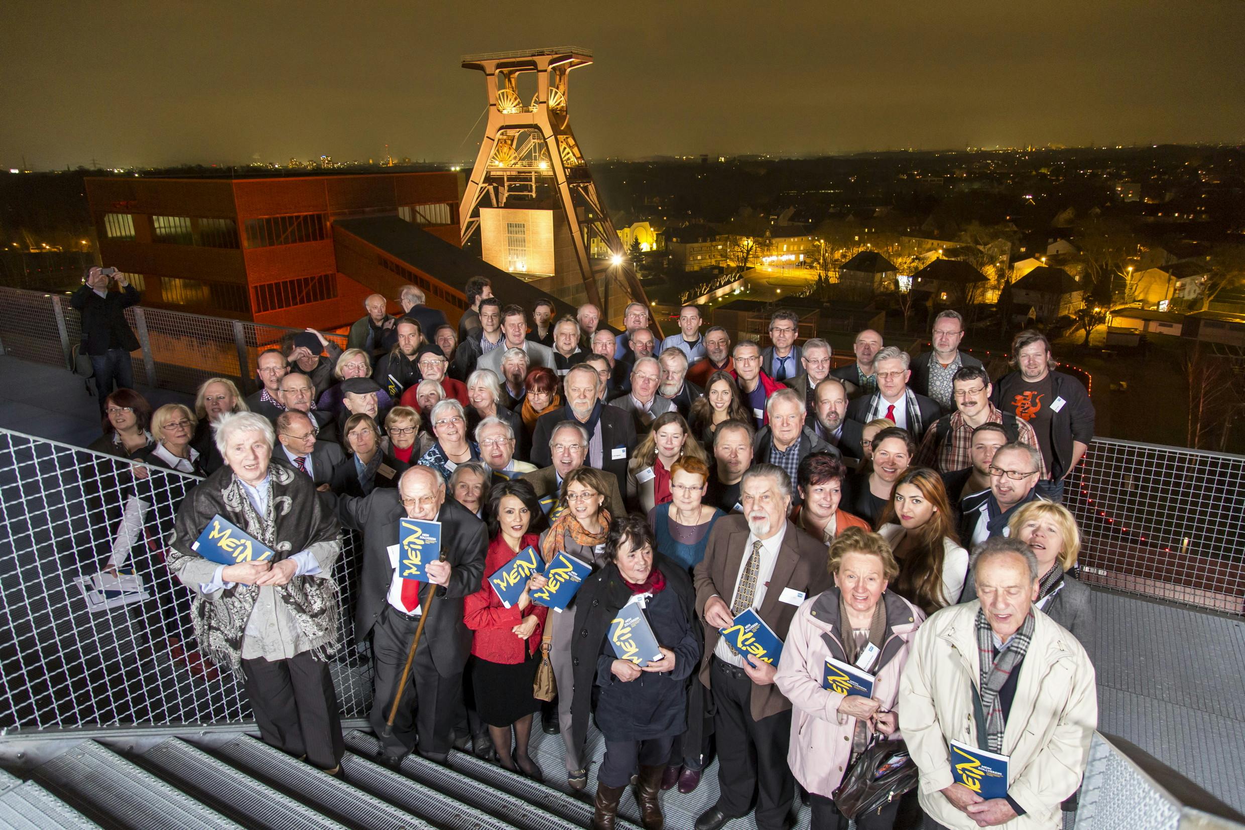 Eine große Gruppe von Menschen steht abends auf einer Aussichtsplattform auf dem Gelände der Zeche Zollverein in Essen. Im Hintergrund ist der berühmte Doppelbock-Förderturm der Schachtanlage 12 beleuchtet und klar zu erkennen. Die Menschen sind unterschiedlichen Alters, viele halten Bücher mit dem Schriftzug Mein Zollverein in den Händen. Einige lächeln direkt in die Kamera. Im Hintergrund erstreckt sich das nächtlich beleuchtete Stadtpanorama mit Häusern und Straßenlaternen. Die Stimmung wirkt feierlich und gemeinschaftlich.