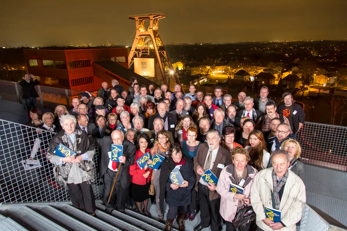 Eine große Gruppe von Menschen steht abends auf einer Aussichtsplattform auf dem Gelände der Zeche Zollverein in Essen. Im Hintergrund ist der berühmte Doppelbock-Förderturm der Schachtanlage 12 beleuchtet und klar zu erkennen. Die Menschen sind unterschiedlichen Alters, viele halten Bücher mit dem Schriftzug Mein Zollverein in den Händen. Einige lächeln direkt in die Kamera. Im Hintergrund erstreckt sich das nächtlich beleuchtete Stadtpanorama mit Häusern und Straßenlaternen. Die Stimmung wirkt feierlich und gemeinschaftlich.