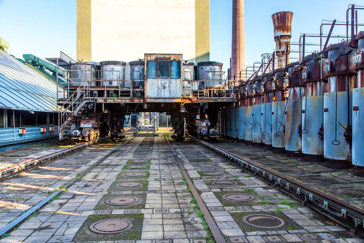 Zentrale Perspektive auf eine historische Koksofenbatterie auf dem Gelände der ehemaligen Kokerei Zollverein in Essen. Im Mittelpunkt steht ein großes, rostiges Maschinenfahrzeug auf Schienen – ein sogenannter Ofenwagen – mit mehreren zylindrischen Behältern und einer verglasten Fahrerkabine mit der Nummer 5. Links und rechts verlaufen Schienen und industrielle Bauwerke aus Metall. Rechts reihen sich nummerierte, blau-graue Ofentüren mit Hebelgriffen aneinander. Der Boden ist mit quadratischen Platten und runden Gusseisenabdeckungen bedeckt, zwischen denen Moos wächst.
