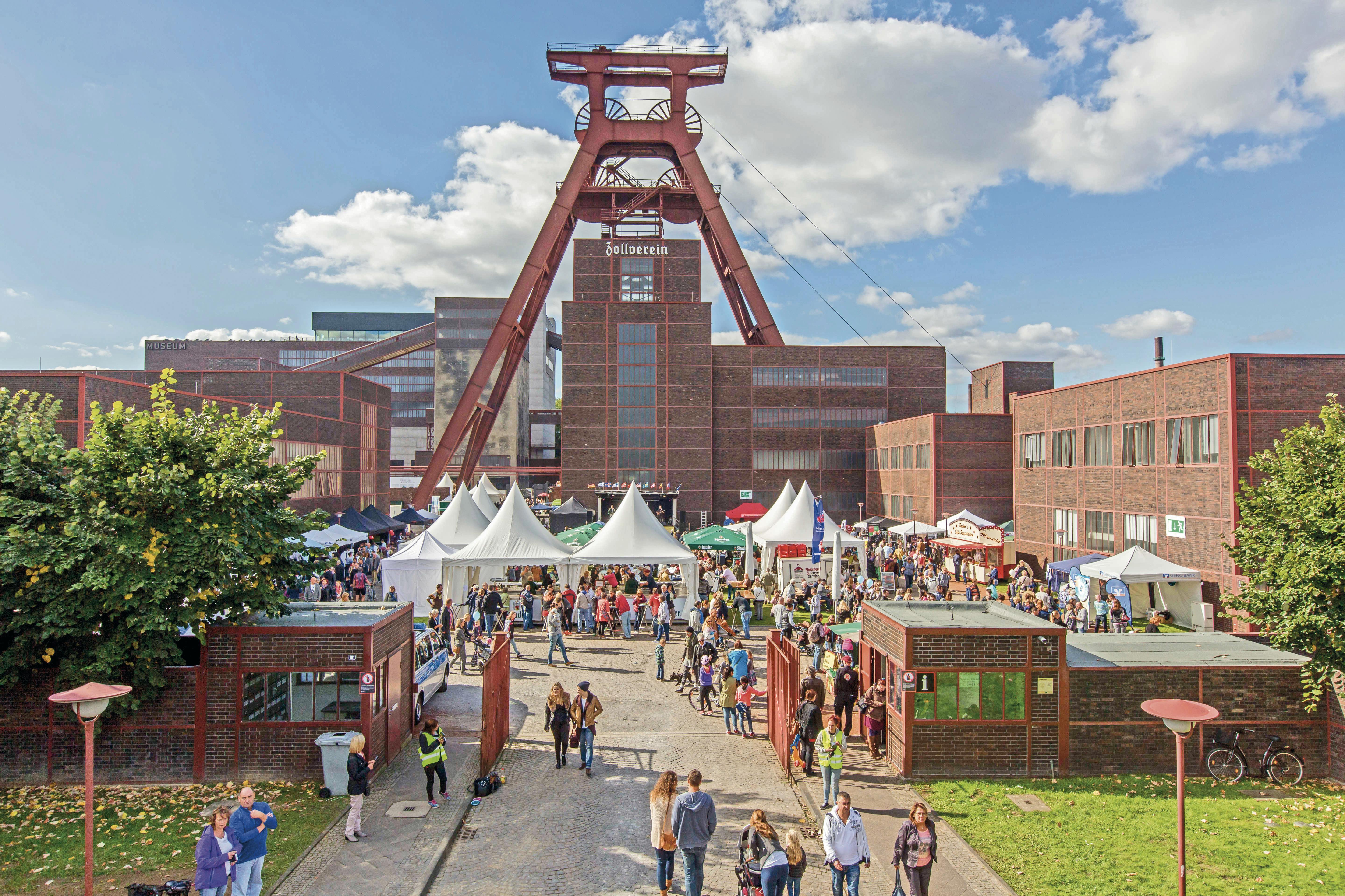 Das Bild zeigt das Gelände des UNESCO-Welterbes Zollverein in Essen an einem sonnigen Tag mit blauem Himmel und weißen Wolken. Im Zentrum steht das markante rote Doppelbock-Fördergerüst von Schacht XII mit dem Schriftzug „Zollverein“. Davor findet ein großes Fest statt: Zahlreiche weiße Pavillons und Verkaufsstände sind aufgebaut, umgeben von vielen Menschen, die durch das Areal schlendern. Die historische rote Backsteinarchitektur der Zeche bildet den Hintergrund. Die Stimmung ist lebendig und einladend.