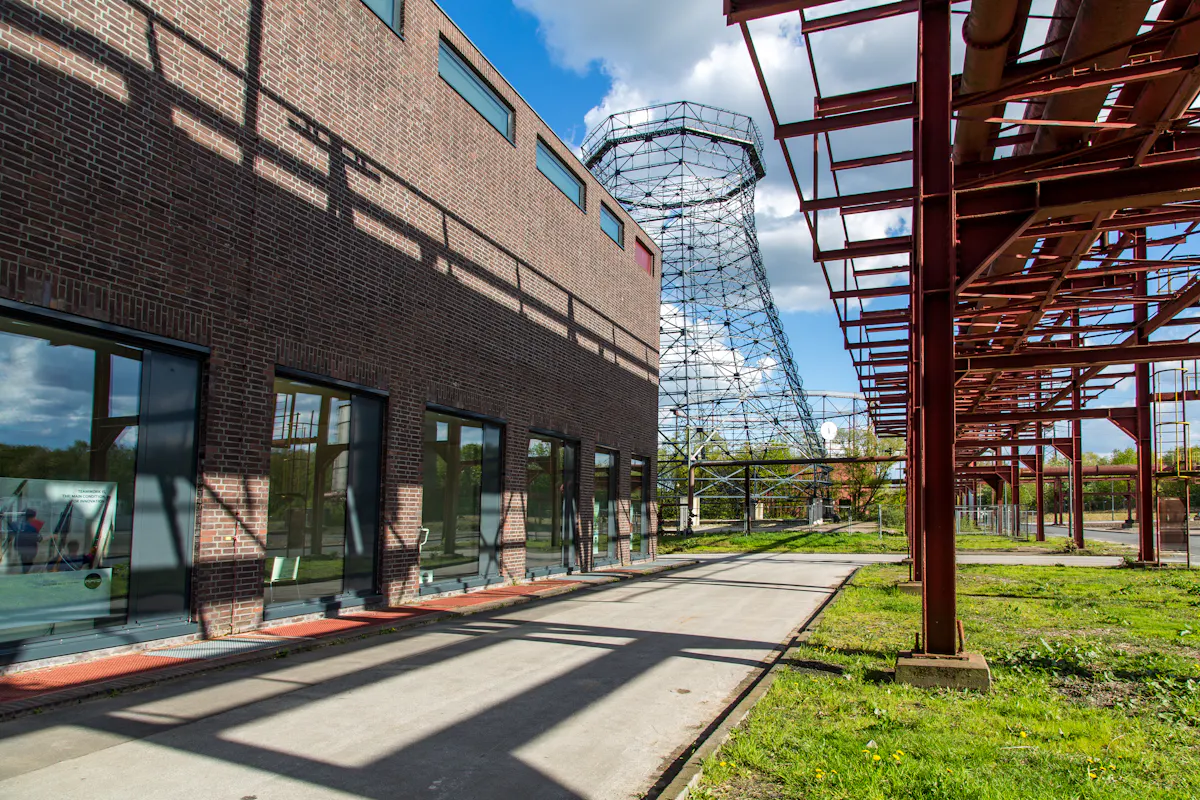 Auf dem Gelände der ehemaligen Kokerei des UNESCO-Welterbes Zollverein in Essen erstreckt sich eine moderne Ziegelfassade mit großen Glasfenstern entlang eines Gehwegs. Rechts daneben ragt ein filigranes Stahlgerüst eines ehemaligen Kühlturms in den Himmel, im Hintergrund ein sonniger, leicht bewölkter Frühlingstag. Über dem Weg verläuft ein rotbraunes Rohrleitungssystem aus Stahlträgern, das teils Schatten auf den Boden und die Gebäudewand wirft. Die Szene zeigt den spannenden Kontrast zwischen industriellem Erbe, zeitgenössischer Architektur und neuem Leben auf dem historischen Gelände.