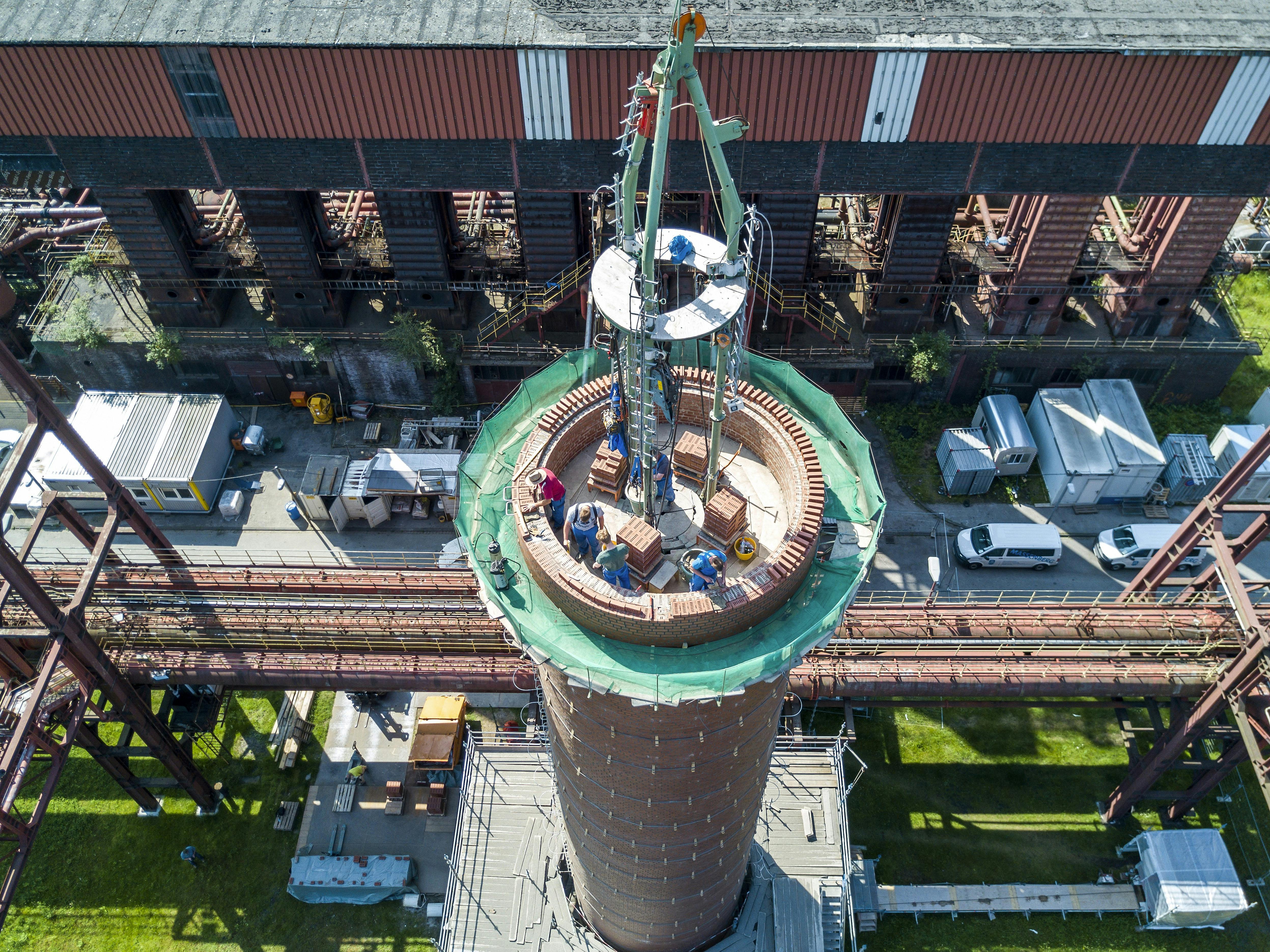 Luftaufnahme einer laufenden Sanierung auf der Kokerei Zollverein in Essen. Im Zentrum des Bildes befindet sich ein großer, runder Schornstein aus rotem Ziegelmauerwerk, der sich mittig ins Bild nach oben erhebt. Auf dem oberen Rand arbeiten mehrere Handwerker unter Sicherungsmaßnahmen am Wiederaufbau des Schornsteins. Ein grünes Schutznetz umgibt die Baustelle. In der Mitte ragt ein montierter Baukran mit Plattform in die Höhe. Im Hintergrund ist ein langgestrecktes Gebäude der ehemaligen Kokerei Zollverein mit markanten roten Streben und schwarzen Öffnungen zu sehen, flankiert von Rohrleitungssystemen und Containern.