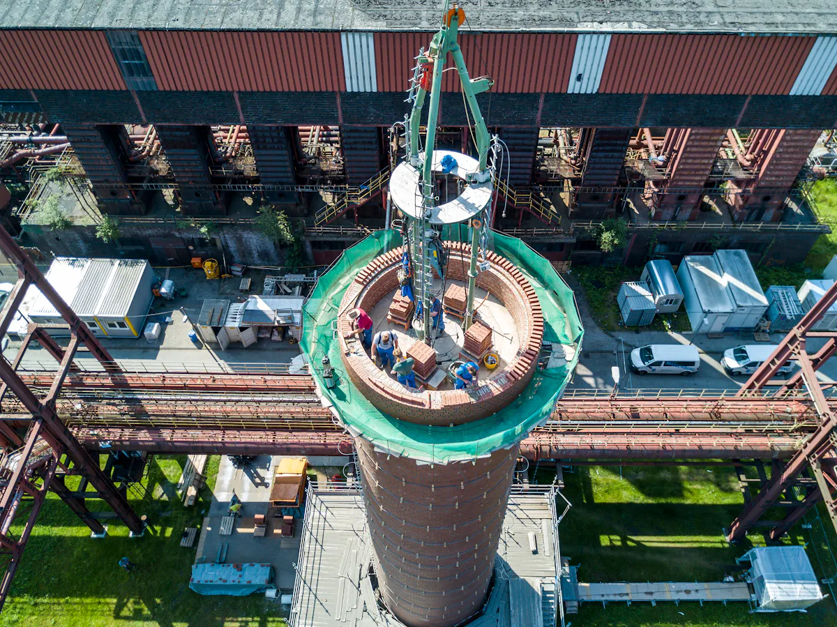 Luftaufnahme einer laufenden Sanierung auf der Kokerei Zollverein in Essen. Im Zentrum des Bildes befindet sich ein großer, runder Schornstein aus rotem Ziegelmauerwerk, der sich mittig ins Bild nach oben erhebt. Auf dem oberen Rand arbeiten mehrere Handwerker unter Sicherungsmaßnahmen am Wiederaufbau des Schornsteins. Ein grünes Schutznetz umgibt die Baustelle. In der Mitte ragt ein montierter Baukran mit Plattform in die Höhe. Im Hintergrund ist ein langgestrecktes Gebäude der ehemaligen Kokerei Zollverein mit markanten roten Streben und schwarzen Öffnungen zu sehen, flankiert von Rohrleitungssystemen und Containern.