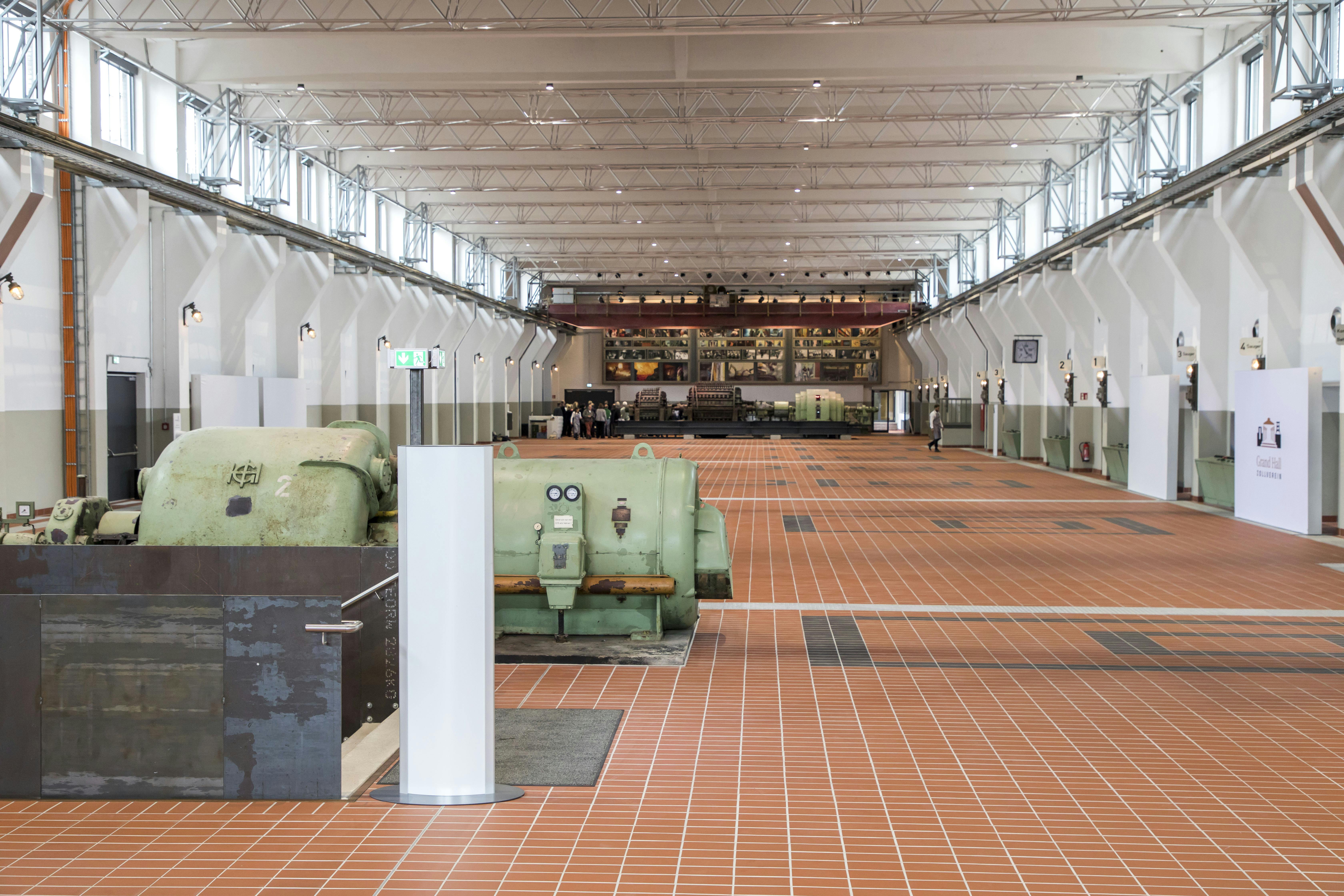 Innenansicht der Grand Hall auf dem UNESCO-Welterbe Zollverein in Essen. Die große Halle mit Oberlichtern und weiß gestrichenen Wänden wurde auf dem historischen Industriegelände modern umgebaut. Der Boden ist mit roten Fliesen ausgelegt und es befinden sich dort alte Maschinenanlagen, darunter große, grün lackierte Generatoren. Die Halle ist lichtdurchflutet und symmetrisch aufgebaut. Im hinteren Bereich sind Menschen zu sehen, die die Größe der Halle verdeutlichen.