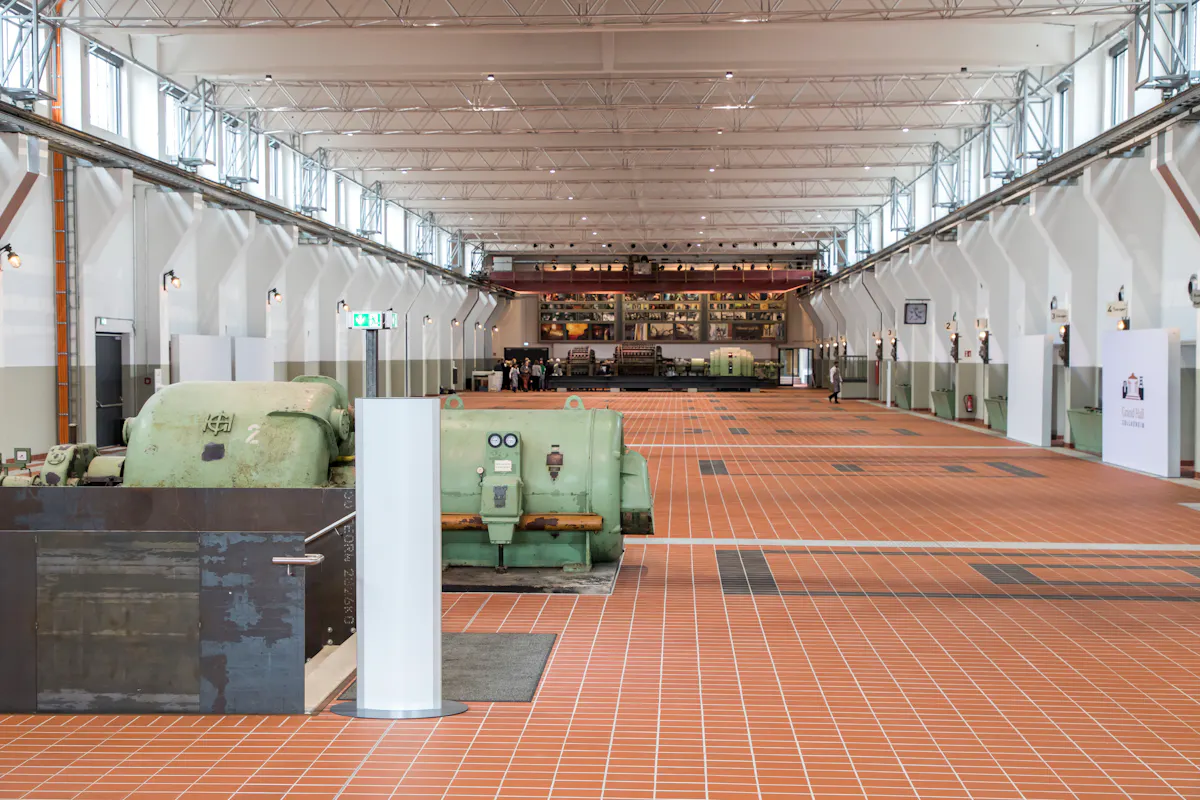 Innenansicht der Grand Hall auf dem UNESCO-Welterbe Zollverein in Essen. Die große Halle mit Oberlichtern und weiß gestrichenen Wänden wurde auf dem historischen Industriegelände modern umgebaut. Der Boden ist mit roten Fliesen ausgelegt und es befinden sich dort alte Maschinenanlagen, darunter große, grün lackierte Generatoren. Die Halle ist lichtdurchflutet und symmetrisch aufgebaut. Im hinteren Bereich sind Menschen zu sehen, die die Größe der Halle verdeutlichen.