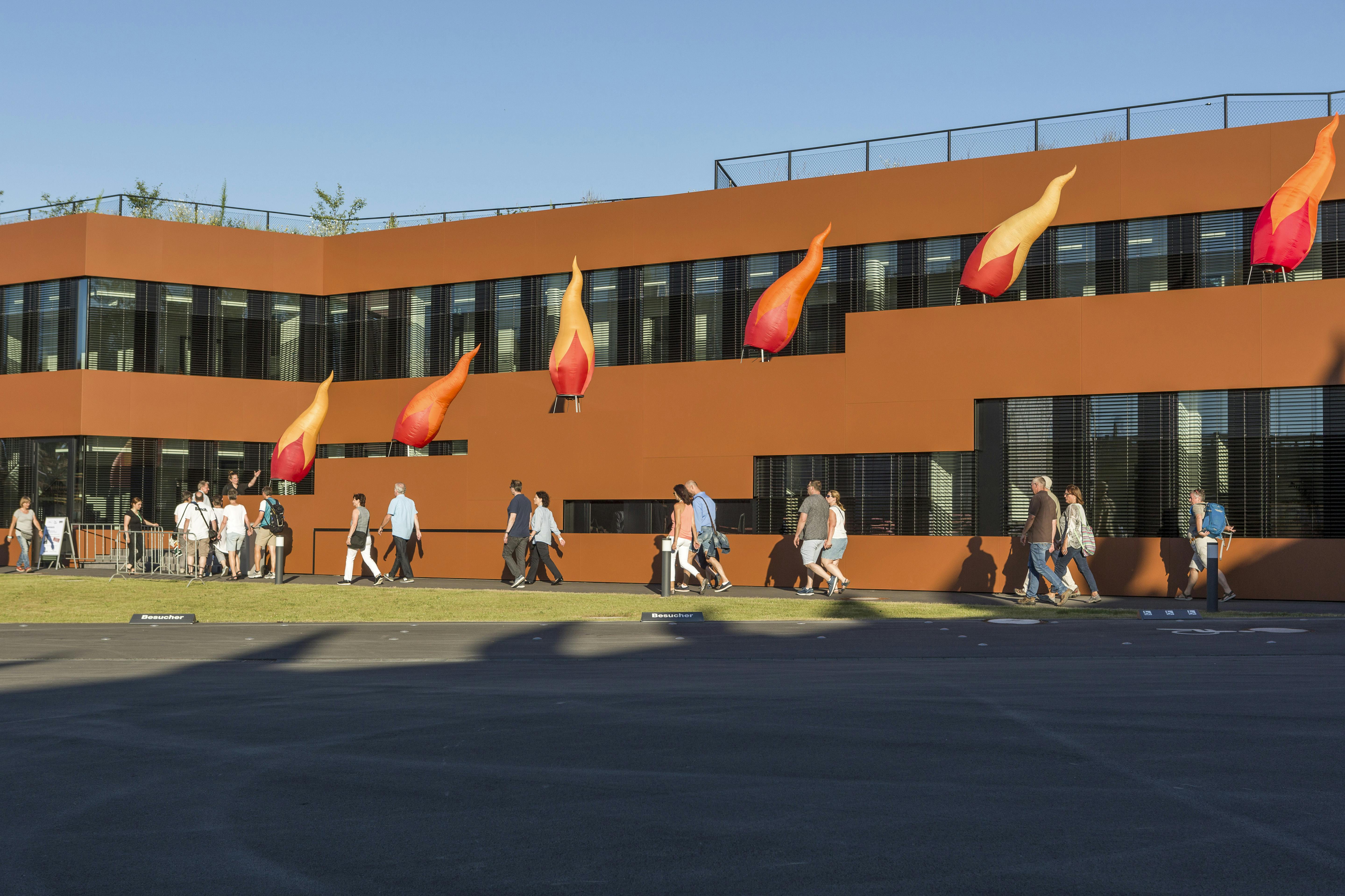 Das Bild zeigt das Gebäude der RAG-Stiftung auf dem Gelände der Kokerei Zollverein in Essen während der Kulturveranstaltung Extraschicht. Die Fassade des modernen, rostrot verkleideten Gebäudes ist mit auffälligen, flammenförmigen Installationen in Orange- und Rottönen geschmückt, die an Feuerzungen erinnern. Vor dem Gebäude gehen zahlreiche Besucherinnen und Besucher entlang, viele in sommerlicher Kleidung, bei sonnigem Wetter und klarem Himmel.