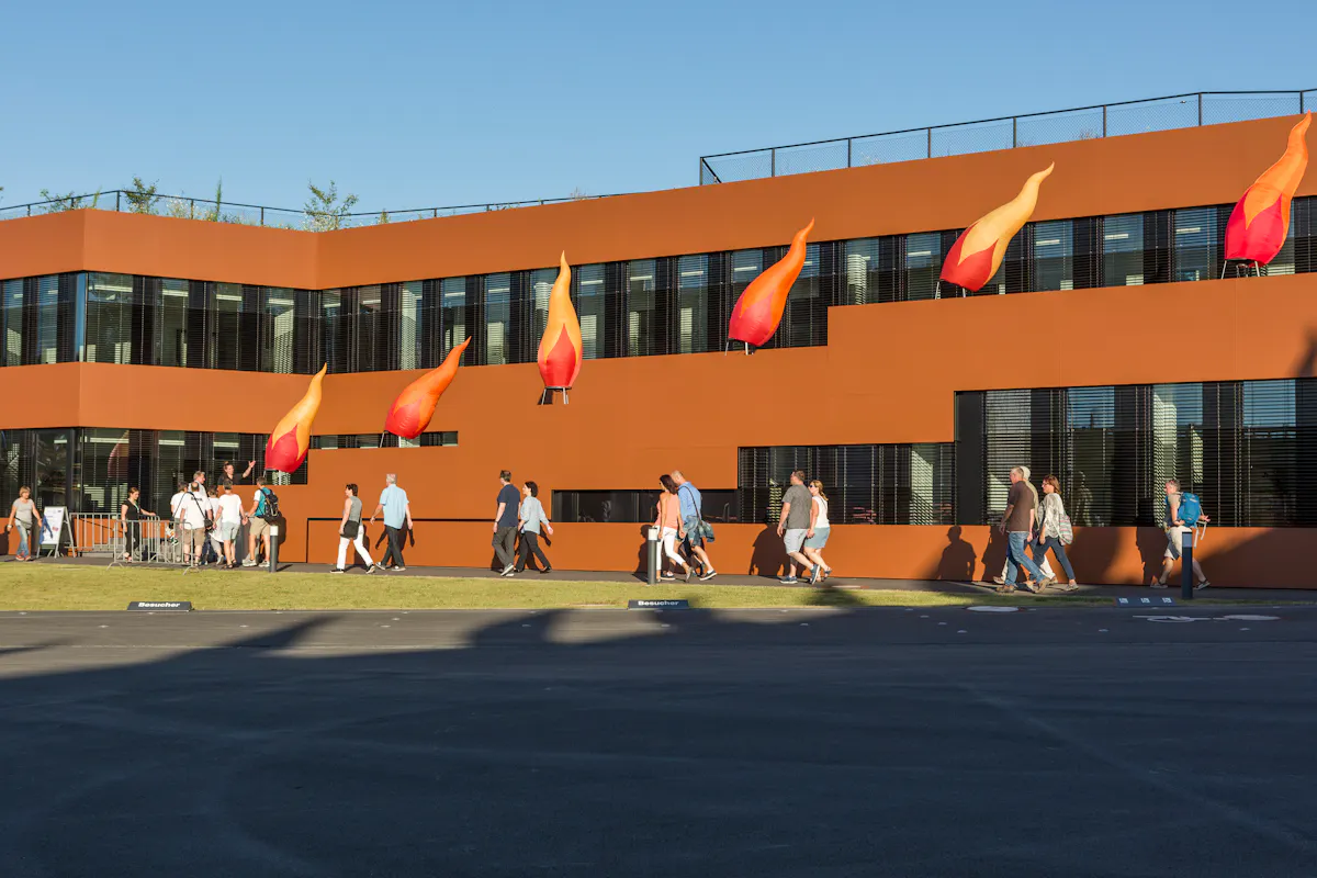 Das Bild zeigt das Gebäude der RAG-Stiftung auf dem Gelände der Kokerei Zollverein in Essen während der Kulturveranstaltung Extraschicht. Die Fassade des modernen, rostrot verkleideten Gebäudes ist mit auffälligen, flammenförmigen Installationen in Orange- und Rottönen geschmückt, die an Feuerzungen erinnern. Vor dem Gebäude gehen zahlreiche Besucherinnen und Besucher entlang, viele in sommerlicher Kleidung, bei sonnigem Wetter und klarem Himmel.