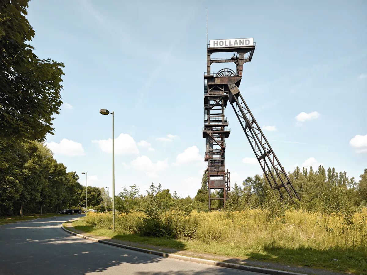 Eine alte Förderturmkonstruktion aus Stahl steht auf einer Wiese am Rand einer Straße. Oben auf dem Turm befindet sich ein Schild mit der Aufschrift HOLLAND. Die Struktur wirkt industriell und rostig und ist von Bäumen und wilder Vegetation umgeben. Links führt eine Straße vorbei, gesäumt von Straßenlaternen und Bäumen. Der Himmel ist blau mit einigen weißen Wolken.