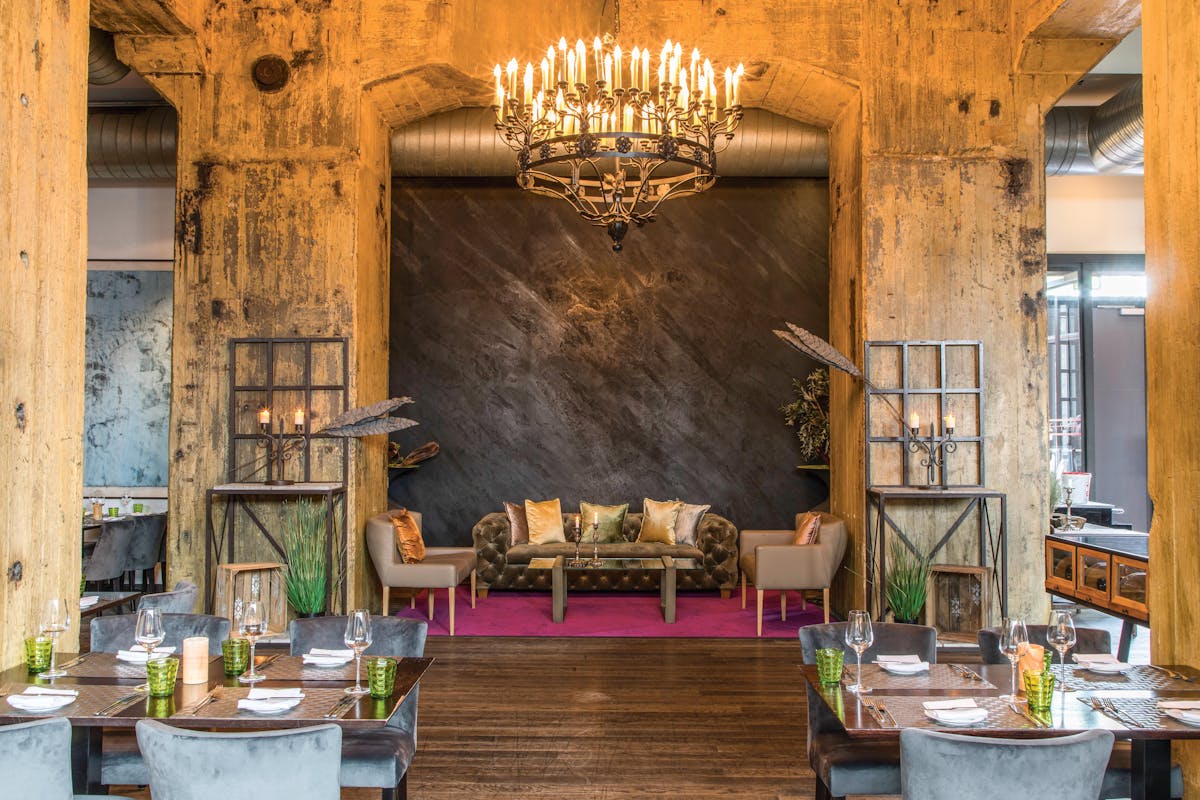 Innenaufnahme des stilvoll eingerichteten Restaurants Casino Zollverein auf dem Gelände der ehemaligen Zeche Zollverein in Essen. Im Mittelpunkt steht eine gemütliche Lounge-Ecke mit einem samtbezogenen Sofa, dekorativen Kissen und zwei Sesseln, eingerahmt von massiven Industrie-Betonpfeilern. Darüber hängt ein großer Kronleuchter mit vielen brennenden Kerzenlampen und sorgt für warmes Licht. Der Boden ist aus dunklem Holz, ein purpurner Teppich hebt den Lounge-Bereich farblich hervor. Im Vordergrund stehen edel gedeckte Restauranttische mit grünen Wassergläsern, Weingläsern und Stoffservietten. Die industrielle Architektur des Gebäudes wird durch dekorative Elemente wie Metallregale, Pflanzen und Kerzen elegant in Szene gesetzt und verbindet Industriegeschichte mit moderner Gastronomie.