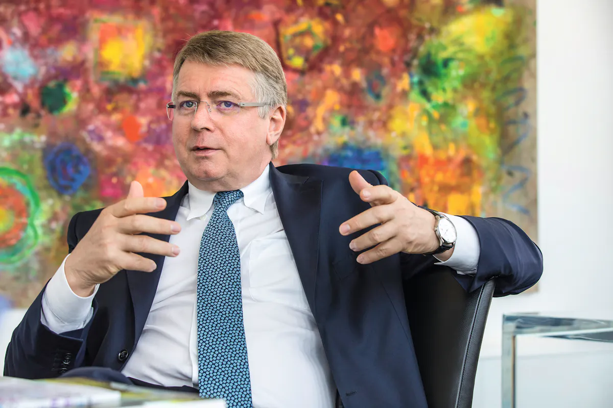 Bernd Tönjes, Vorsitzender des Vorstandes der RAG-Stiftung, sitzt in einem modernen Bürointerieur und spricht gestikulierend mit ausdrucksstarker Körpersprache. Er trägt einen dunklen Anzug, ein weißes Hemd und eine blau-gemusterte Krawatte. Hinter ihm hängt ein farbenfrohes, abstraktes Gemälde. Das Bild vermittelt eine angeregte Gesprächssituation.