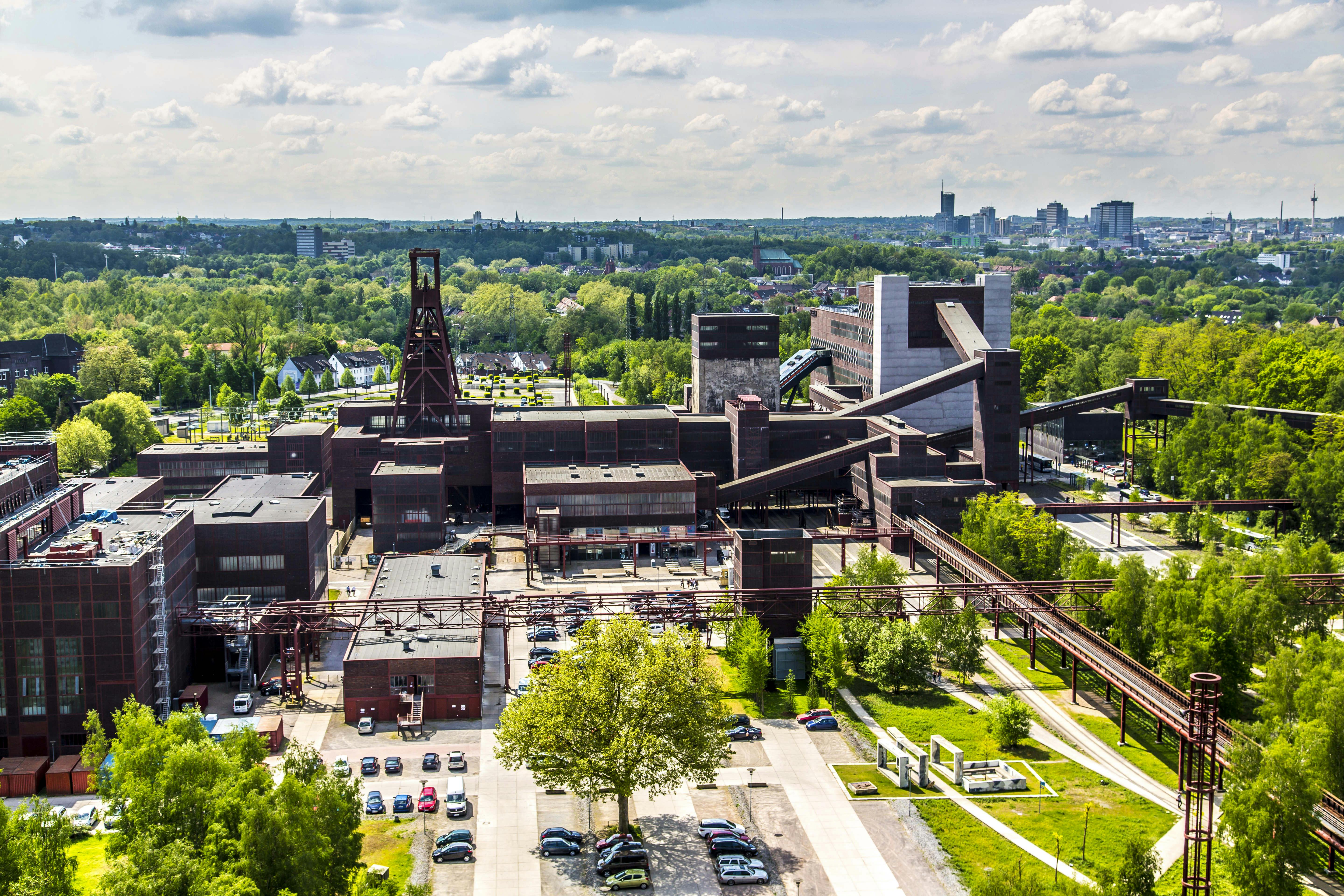 Luftaufnahme des UNESCO-Welterbes Zollverein in Essen an einem sonnigen Tag. Im Zentrum des Bildes steht das charakteristische rote Doppelbock-Fördergerüst von Schacht XII, umgeben von symmetrisch angeordneten Backsteingebäuden mit dunkler Fassade und roten Fensterrahmen. Mehrere ehemalige Industrieanlagen, Förderbänder und Rohrleitungen verlaufen über das Gelände, das heute durch begrünte Wege, Bäume und Parkplätze durchzogen ist. Im Hintergrund ist die grüne Stadtlandschaft des Ruhrgebiets mit einzelnen Hochhäusern und Kirchtürmen zu sehen.