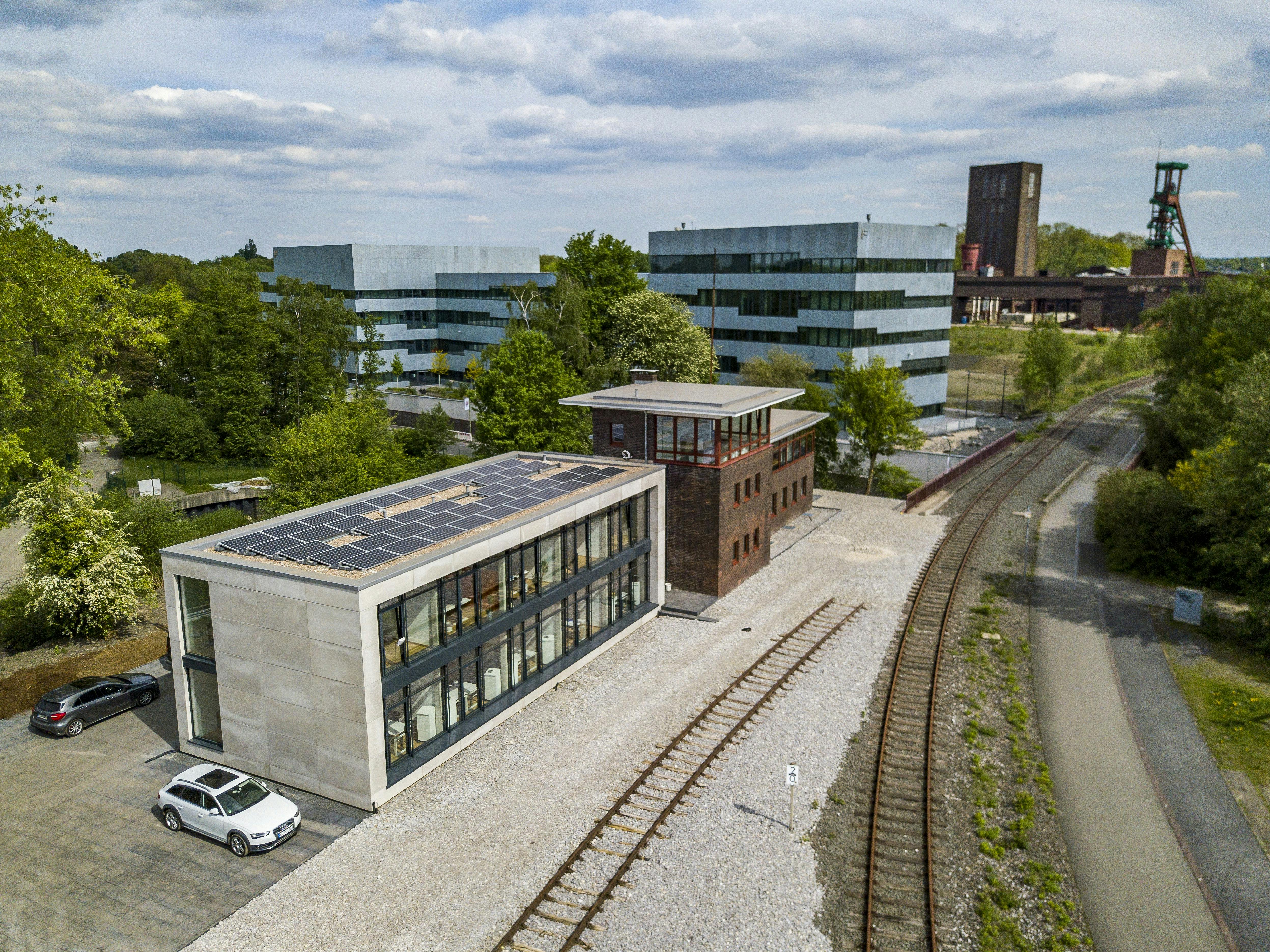 Das Bild zeigt eine moderne Szene auf dem Gelände der Zeche Zollverein in Essen. Im Vordergrund befindet sich das Stellwerk Kokerei – ein saniertes, rechteckiges Gebäude aus Backstein mit großen Fenstern und einem modernen Anbau mit Solarpaneelen auf dem Dach. Daneben verlaufen stillgelegte Bahngleise auf hellem Schotter. Im Hintergrund stehen die markanten, silbrig-blau verkleideten Gebäude der Folkwang Universität der Künste. Dahinter ragt der Förderturm der Schachtanlage Zollverein 1/2/8 in den Himmel. Die Umgebung ist grün und von Bäumen gesäumt, bei bewölktem Himmel.