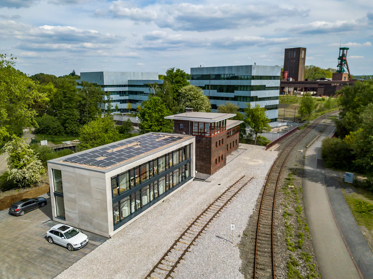 Das Bild zeigt eine moderne Szene auf dem Gelände der Zeche Zollverein in Essen. Im Vordergrund befindet sich das Stellwerk Kokerei – ein saniertes, rechteckiges Gebäude aus Backstein mit großen Fenstern und einem modernen Anbau mit Solarpaneelen auf dem Dach. Daneben verlaufen stillgelegte Bahngleise auf hellem Schotter. Im Hintergrund stehen die markanten, silbrig-blau verkleideten Gebäude der Folkwang Universität der Künste. Dahinter ragt der Förderturm der Schachtanlage Zollverein 1/2/8 in den Himmel. Die Umgebung ist grün und von Bäumen gesäumt, bei bewölktem Himmel.