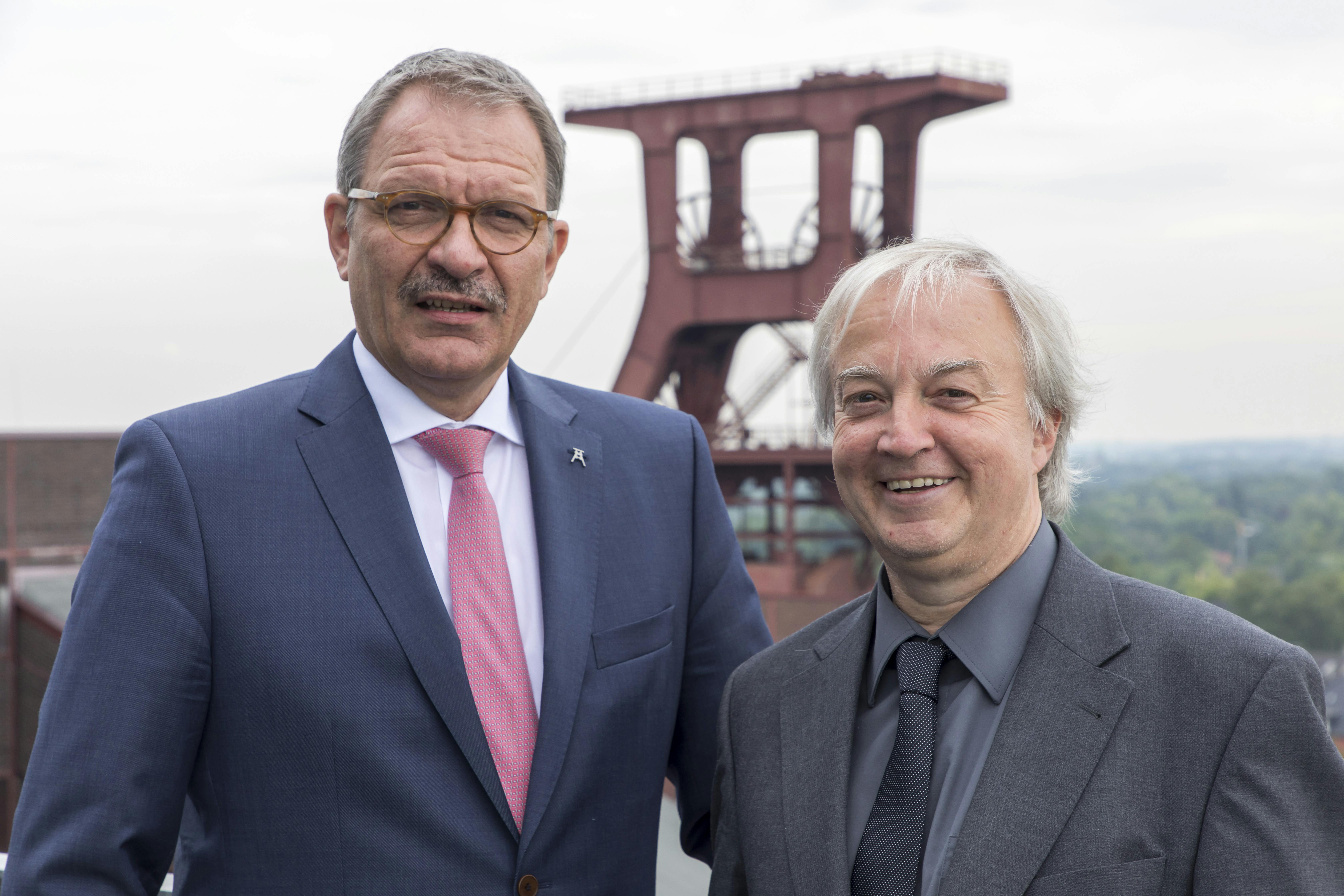 or dem berühmten Doppelbock-Fördergerüst der Zeche Zollverein in Essen stehen zwei Männer im Anzug. Links steht Prof. Hans-Peter Noll, Vorstandsvorsitzender der Stiftung Zollverein, im blauen Anzug mit rosa Krawatte und Brille. Rechts neben ihm steht Prof. Heinrich Theodor Grütter, ebenfalls im Vorstand der Stiftung und Direktor des Ruhr Museums, mit grauem Haar, dunkelgrauem Anzug und einem freundlichen Lächeln. Der Hintergrund zeigt das historische Industriedenkmal und einen weiten Blick ins grüne Ruhrgebiet.