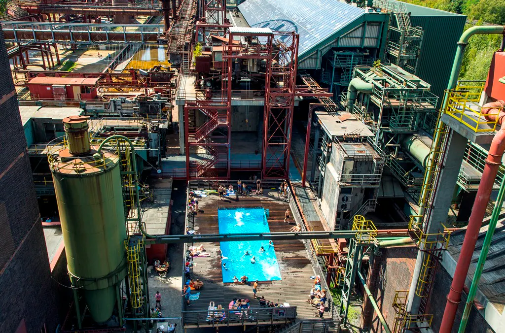 Luftaufnahme des Werksschwimmbads auf dem Gelände der ehemaligen Kokerei Zollverein in Essen. Inmitten einer beeindruckenden Kulisse aus rostigem Stahl, Industrieanlagen, Rohren und Förderbändern liegt ein leuchtend blaues Schwimmbecken mit einem umgebenden Holzdeck. Menschen baden im Pool oder entspannen auf Liegen rundherum. Die umgebenden Industrieelemente – hohe Rohre, Stahltreppen und Maschinengehäuse – kontrastieren stark mit der sommerlich-leichten Atmosphäre rund um das Becken.