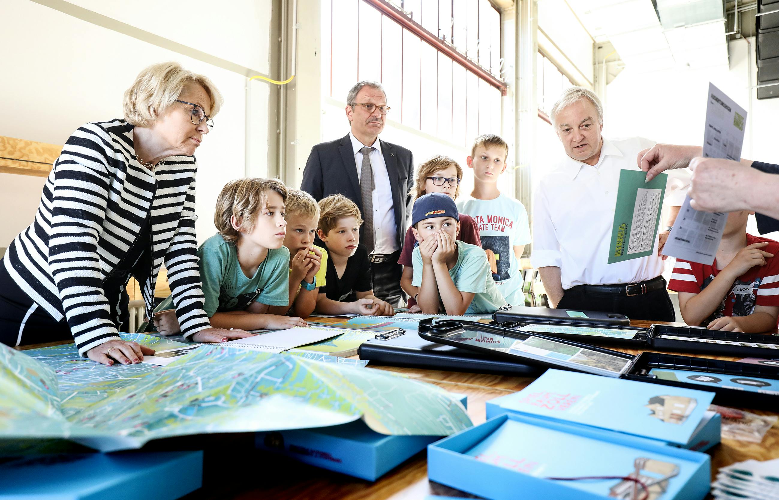 Eine Gruppe von Kindern und Erwachsenen steht um einen großen Tisch, auf dem Karten, Hefte und Bildungsmaterialien ausgelegt sind. Links im Bild lehnt sich Bärbel Bergerhoff-Wodopia, Mitglied des Vorstands der RAG-Stiftung, aufmerksam über die Tischkante. Rechts von ihr stehen mehrere Kinder, die gespannt den Erklärungen folgen. Im Hintergrund stehen Prof. Noll und Prof. Grütter. Ein Erwachsener hält erklärend Arbeitsblätter in der Hand. Die Szene spielt sich in einem hellen Raum mit hohen Fenstern ab und zeigt ein Bildungsangebot für Kinder im Rahmen eines Natur- und Erlebnisprogramms auf dem Welterbe Zollverein.
