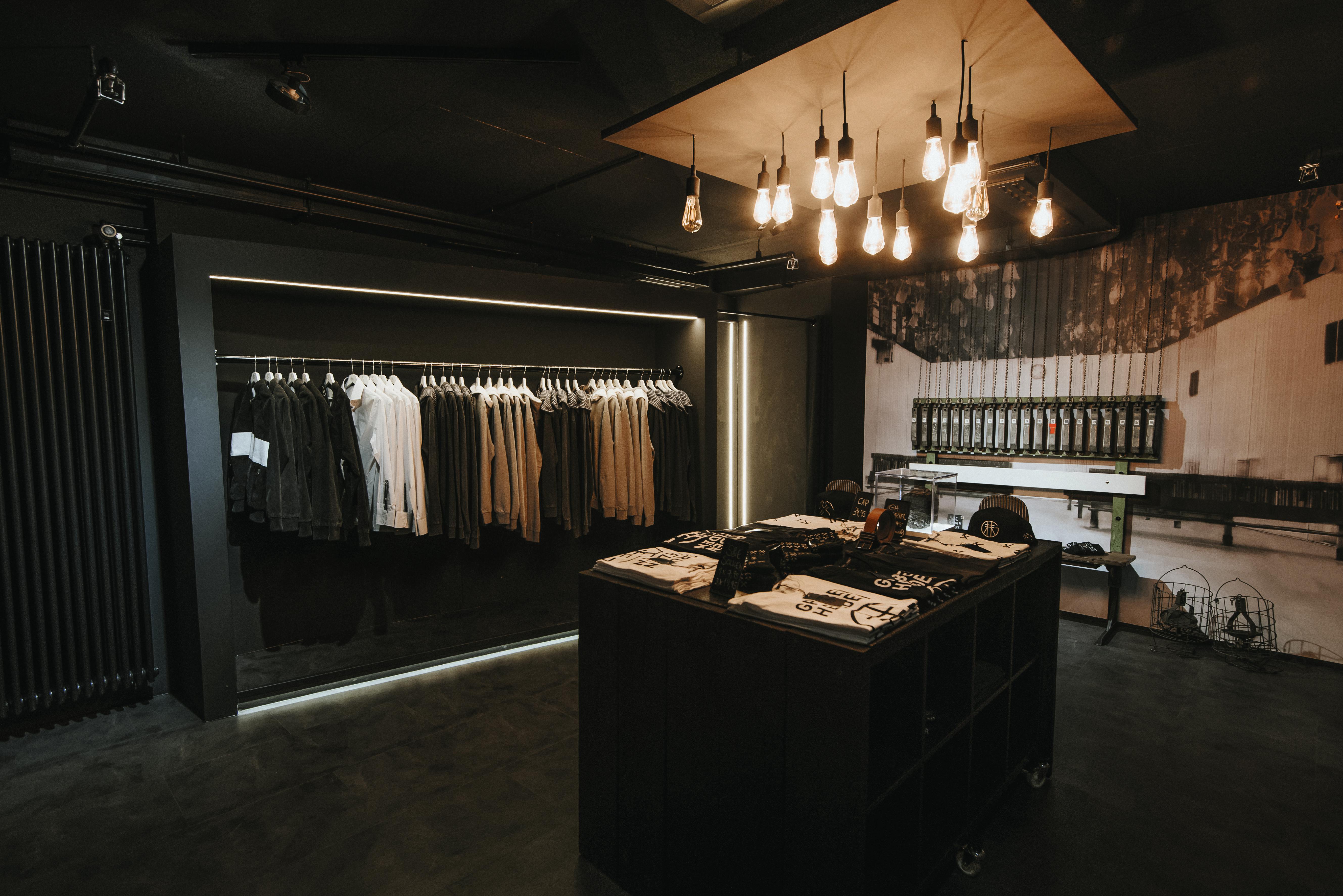 Im Grubenhelden-Store auf dem UNESCO-Welterbe Zollverein in Essen herrscht eine minimalistische, dunkle Atmosphäre mit gezielter Beleuchtung. Links hängen schwarze, weiße und beige Kleidungsstücke ordentlich auf einer Kleiderstange, darunter Hoodies, Hemden und Jacken – alle im schlichten, bergbauinspirierten Design. In der Mitte steht ein schwarzer Tisch mit neatly gefalteten T-Shirts, Caps und Accessoires, viele davon tragen das Grubenhelden-Logo oder typische Bergbausymbole. Im Hintergrund dominiert ein großes Wandbild einer historischen Kaue – einer Umkleidehalle mit herabhängenden Körben aus dem Bergbau – und verleiht dem Raum einen authentischen Bezug zur Geschichte der Kumpel. Die Einrichtung und das Lichtkonzept betonen den industriellen Charme des Ruhrgebiets.