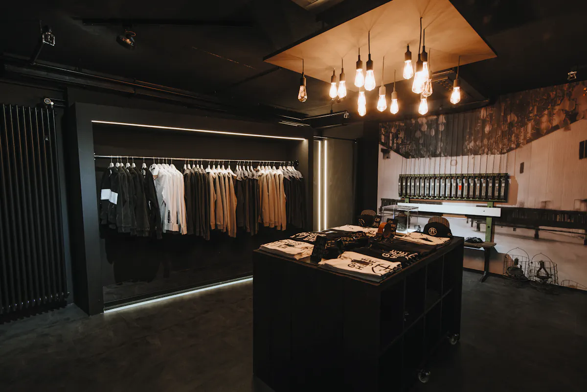 Im Grubenhelden-Store auf dem UNESCO-Welterbe Zollverein in Essen herrscht eine minimalistische, dunkle Atmosphäre mit gezielter Beleuchtung. Links hängen schwarze, weiße und beige Kleidungsstücke ordentlich auf einer Kleiderstange, darunter Hoodies, Hemden und Jacken – alle im schlichten, bergbauinspirierten Design. In der Mitte steht ein schwarzer Tisch mit neatly gefalteten T-Shirts, Caps und Accessoires, viele davon tragen das Grubenhelden-Logo oder typische Bergbausymbole. Im Hintergrund dominiert ein großes Wandbild einer historischen Kaue – einer Umkleidehalle mit herabhängenden Körben aus dem Bergbau – und verleiht dem Raum einen authentischen Bezug zur Geschichte der Kumpel. Die Einrichtung und das Lichtkonzept betonen den industriellen Charme des Ruhrgebiets.