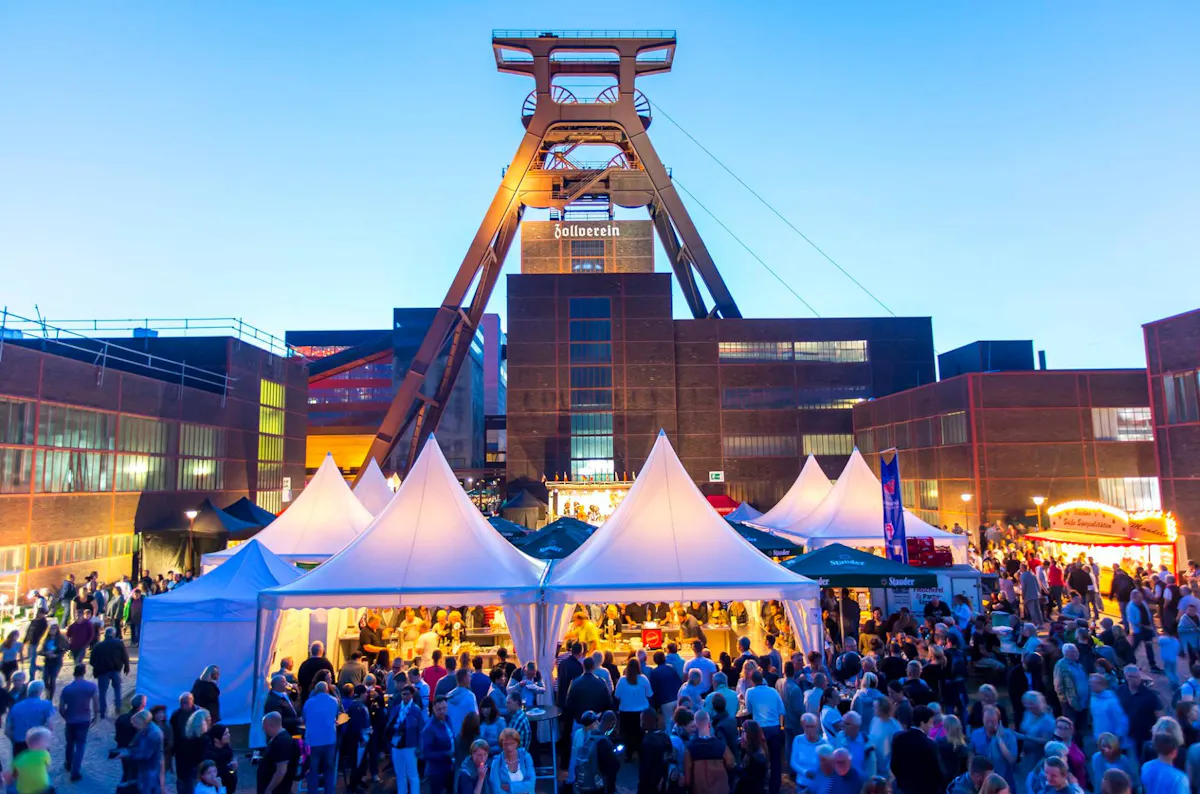 Auf dem Vorplatz des beleuchteten Doppelbock-Fördergerüsts der Zeche Zollverein in Essen findet bei einbrechender Dunkelheit ein großes Fest statt. Weiße Zelte mit spitzen Dächern reihen sich dicht aneinander. Darunter befinden sich Getränkestände und Essensbuden, an denen zahlreiche Besucherinnen und Besucher stehen. Die Szene ist farbenfroh, lebendig und von warmem Licht erfüllt. Die Menschen genießen die Atmosphäre eines Sommerabends vor der eindrucksvollen Industriekulisse des UNESCO-Welterbes.