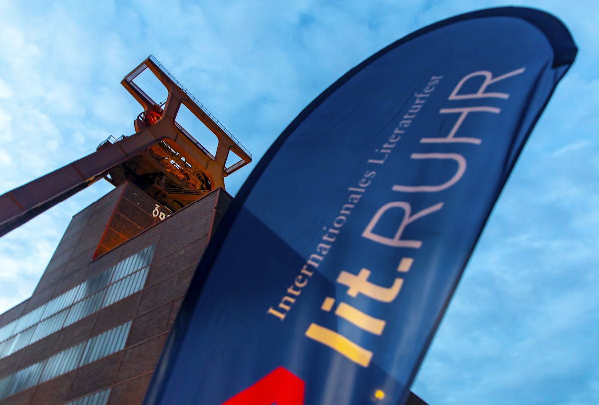 Im Vordergrund weht eine blaue Festivalfahne mit der Aufschrift lit.RUHR – Internationales Literaturfest. Im Hintergrund ragt das markante Fördergerüst der Zeche Zollverein in Essen empor, angestrahlt vom warmen Licht in der Abenddämmerung. Die dunkle Backsteinfassade des Schachtgebäudes bildet einen starken Kontrast zum hellen Himmel. Die Aufnahme symbolisiert die Verbindung von Industriekultur und zeitgenössischer Literatur, wie sie auf dem Welterbe Zollverein regelmäßig erlebbar ist.