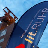 Im Vordergrund weht eine blaue Festivalfahne mit der Aufschrift lit.RUHR – Internationales Literaturfest. Im Hintergrund ragt das markante Fördergerüst der Zeche Zollverein in Essen empor, angestrahlt vom warmen Licht in der Abenddämmerung. Die dunkle Backsteinfassade des Schachtgebäudes bildet einen starken Kontrast zum hellen Himmel. Die Aufnahme symbolisiert die Verbindung von Industriekultur und zeitgenössischer Literatur, wie sie auf dem Welterbe Zollverein regelmäßig erlebbar ist.