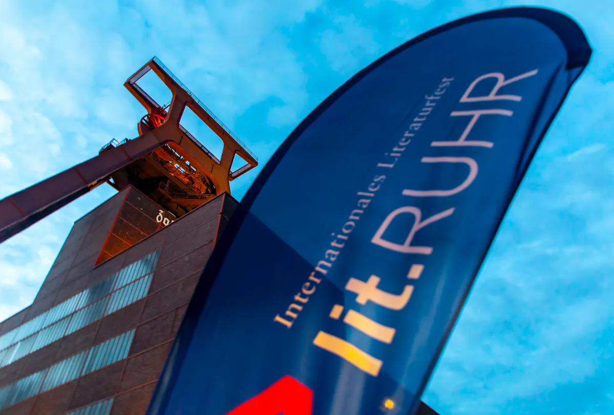 Im Vordergrund weht eine blaue Festivalfahne mit der Aufschrift lit.RUHR – Internationales Literaturfest. Im Hintergrund ragt das markante Fördergerüst der Zeche Zollverein in Essen empor, angestrahlt vom warmen Licht in der Abenddämmerung. Die dunkle Backsteinfassade des Schachtgebäudes bildet einen starken Kontrast zum hellen Himmel. Die Aufnahme symbolisiert die Verbindung von Industriekultur und zeitgenössischer Literatur, wie sie auf dem Welterbe Zollverein regelmäßig erlebbar ist.