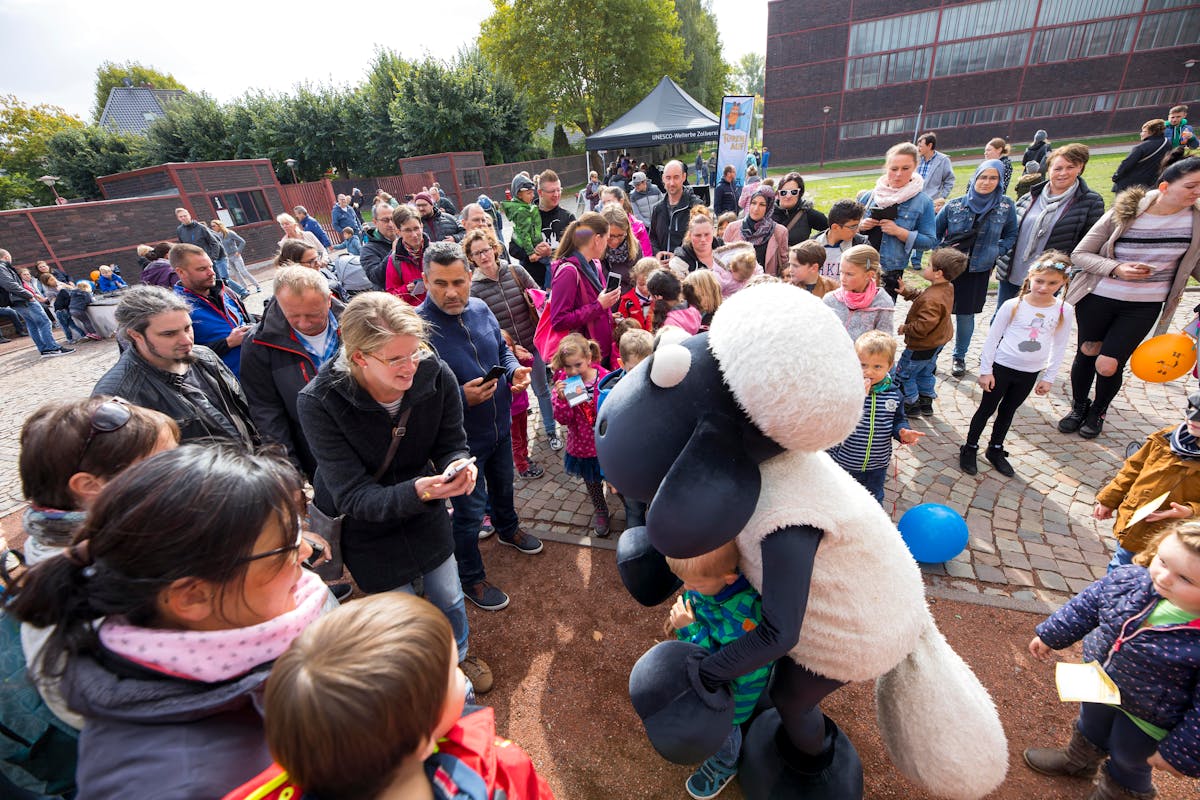 Auf dem Außengelände des UNESCO-Welterbes Zollverein versammelt sich eine große Gruppe von Familien mit Kindern zum beliebten Maustag. Im Mittelpunkt steht die bekannte Figur „Shaun das Schaf“ in einem lebensgroßen Kostüm, das von einem Darsteller getragen wird. Kinder umringen die Figur begeistert und ein Junge wird von Shaun umarmt, während die Eltern Fotos machen. Im Hintergrund stehen weitere Besucher sowie ein schwarzes Zelt mit der Aufschrift „UNESCO-Welterbe Zollverein“. Die Atmosphäre ist fröhlich und lebendig – ein buntes Familienfest mit vielen kleinen Entdeckern.