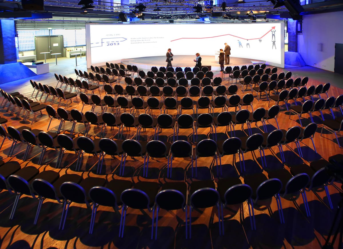 Innenaufnahme des Oktogons auf dem UNESCO-Welterbe Zollverein, einem Veranstaltungsort im ehemaligen Gasometer der Zeche. Der Raum ist für eine Präsentation oder Tagung vorbereitet: Mehrere Reihen schwarzer Stühle sind in einem Halbrund auf den Holzboden ausgerichtet und blicken auf eine große, weiß beleuchtete Projektionswand mit Grafiken und Texten. Einige Personen stehen noch vor der Bühne und bereiten sich auf den Beginn vor. Die Halle ist modern ausgeleuchtet, mit sichtbarer Bühnentechnik und einer klaren industriellen Ästhetik. Das Oktogon dient hier als stilvolle Kulisse für eine professionelle Veranstaltung.