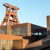 Ansicht des ikonischen Doppelbock-Fördergerüsts der Zeche Zollverein in Essen bei strahlendem Sonnenschein. Im Vordergrund sind die klar strukturierten Backsteinbauten der ehemaligen Schachtanlage XII mit ihren großen Fensterbändern zu sehen. Die markante rote Stahlkonstruktion des Förderturms ragt über die Gebäude hinaus in den hellblauen Himmel.