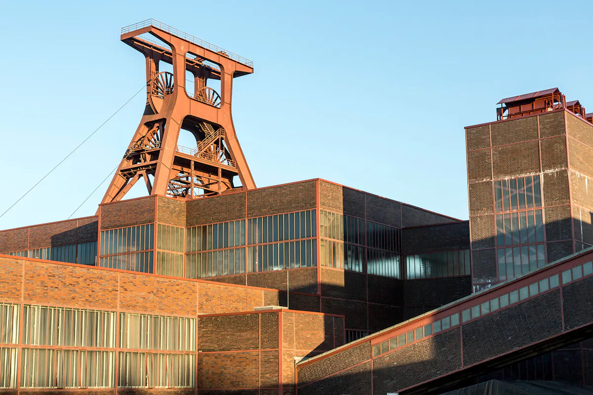 Ansicht des ikonischen Doppelbock-Fördergerüsts der Zeche Zollverein in Essen bei strahlendem Sonnenschein. Im Vordergrund sind die klar strukturierten Backsteinbauten der ehemaligen Schachtanlage XII mit ihren großen Fensterbändern zu sehen. Die markante rote Stahlkonstruktion des Förderturms ragt über die Gebäude hinaus in den hellblauen Himmel.