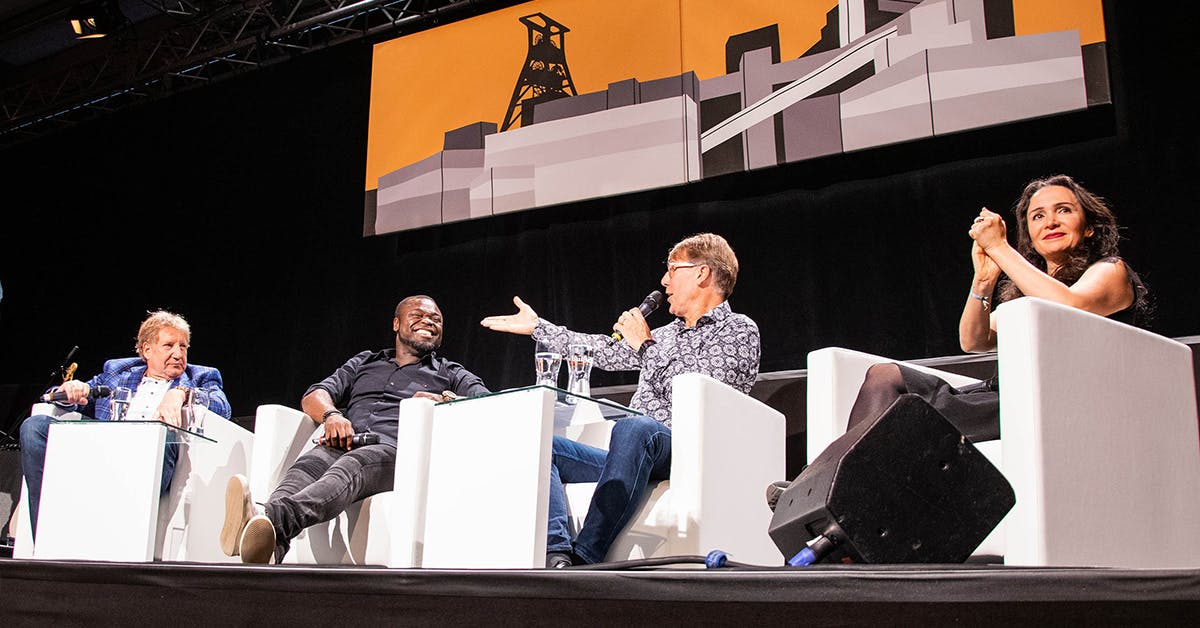 Auf der Bühne des Veranstaltungsformats #halbzwölf auf dem UNESCO-Welterbe Zollverein sitzen vier bekannte Persönlichkeiten von links nach rechts: Moderator Theo Spanke, Ex-Fußballprofi Gerald Asamoah, Sportmoderator Peter Großmann und Kabarettistin Senay Duzcu. Alle sitzen in weißen Sesseln vor schwarzem Hintergrund. Peter Großmann spricht gestikulierend ins Mikrofon, während Gerald Asamoah lacht und Theo Spanke aufmerksam zuhört. Senay Duzcu sitzt am rechten Rand der Bühne und applaudiert mit einem Lächeln. Über der Bühne hängt ein stilisiertes Banner der Zeche Zollverein auf orangefarbenem Hintergrund – ein Symbol für den Veranstaltungsort und das industrielle Erbe.