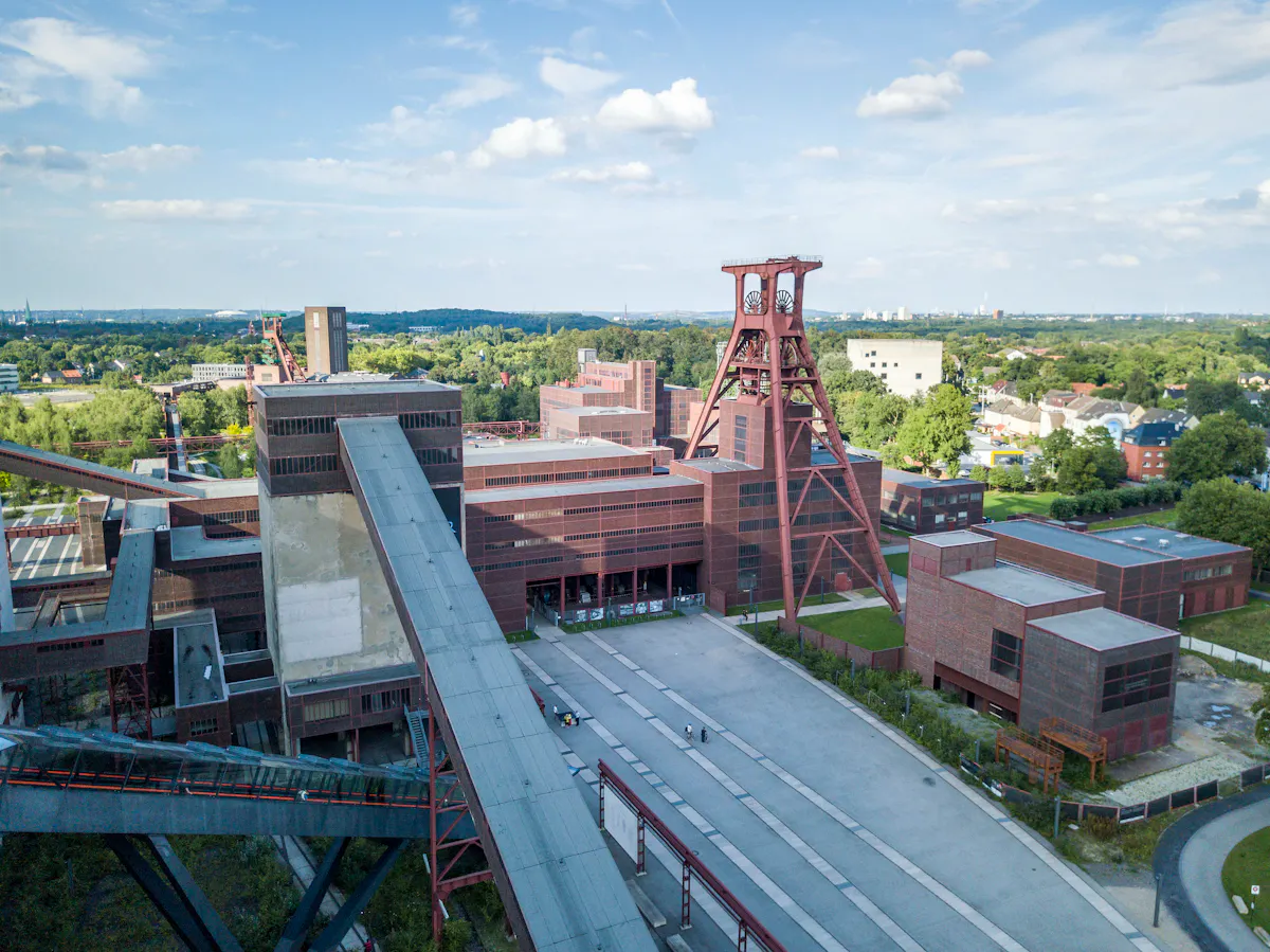 Luftaufnahme des UNESCO-Welterbes Zollverein in Essen bei Tageslicht. Im Zentrum steht das markante Doppelbock-Fördergerüst auf Schacht XII, das mit seiner roten Stahlkonstruktion als architektonisches Wahrzeichen des Ruhrgebiets gilt. Umgeben ist es von symmetrisch angeordneten Backsteingebäuden im Bauhaus-Stil. Förderbänder verlaufen wie Adern über das Gelände. Im Hintergrund erstreckt sich das grüne Ruhrgebiet mit Bäumen, Häusern und dem Horizont.