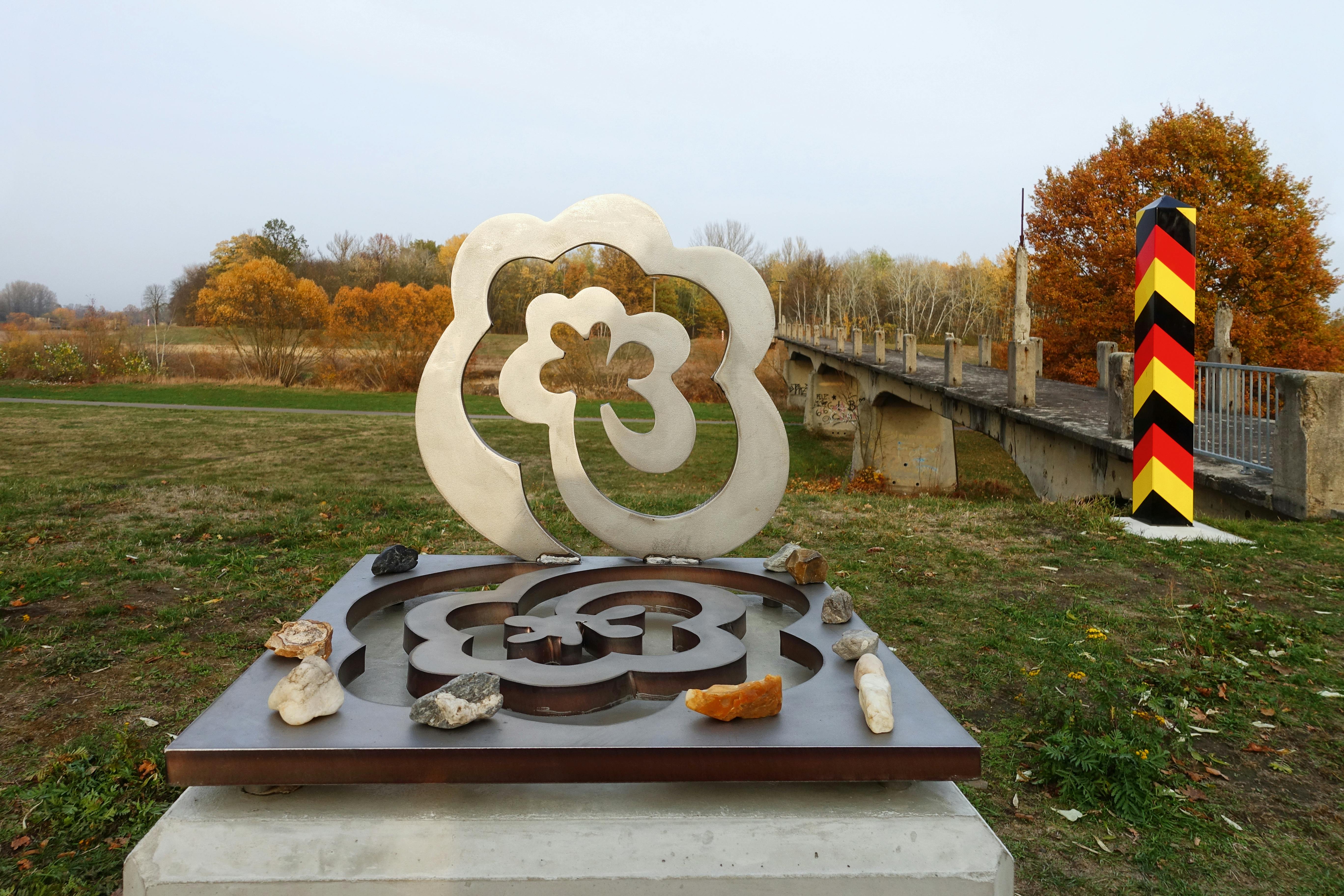 Im Zentrum des Bildes steht eine Metallskulptur auf einem Betonsockel in einer offenen Landschaft mit herbstlicher Färbung. Die Skulptur trägt den Titel Grenzrosen und stammt vom Künstler Thomas Rother. Sie zeigt zwei ineinandergreifende stilisierte Rosenformen, die in positiver und negativer Form gearbeitet sind: Die untere Ebene ist eine eingelassene, vertiefte Negativform einer Rose, aus der eine plastische Positivform in gleicher Kontur emporwächst. Diese steht wie ein Rahmen frei in der Luft, wodurch sich ein harmonischer Durchblick zur Landschaft ergibt. Auf dem Sockel sind kleine Steine und Fundstücke abgelegt – als Zeichen individueller Erinnerung oder Gedenken, ähnlich wie auf jüdischen Gräbern. Im Hintergrund rechts führt eine alte Betonbrücke über den Fluss Neiße – sie war früher Grenzübergang zwischen Deutschland und Polen. Daneben steht ein auffällig bunt gestrichener Grenzpfahl in den Farben Schwarz-Rot-Gold, der die deutsch-polnische Grenze markiert. Die Landschaft im Hintergrund ist flach, mit Bäumen in herbstlichem Orange und Gelb. Der Himmel ist lichtgrau, ruhig, fast kontemplativ.