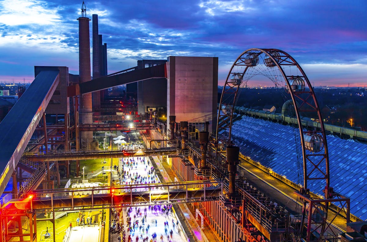 Blick bei Dämmerung auf das beleuchtete Winterdorf der Kokerei Zollverein in Essen. In der Mitte des Bildes verläuft eine lange Eisbahn durch die monumentale Industriekulisse der ehemaligen Kokerei. Die Eisfläche ist von buntem Licht in Blau, Lila und Gelb illuminiert, während viele Menschen dort Schlittschuh laufen. Rechts erhebt sich ein beleuchtetes, großes Riesenrad ohne Bewegung. Die stillgelegten Industrieanlagen – Hochöfen, Rohrleitungen und Hallen – bilden einen imposanten Kontrast zur fröhlichen Winteratmosphäre. Die untergehende Sonne taucht den Himmel in dramatische Blau- und Orangetöne.