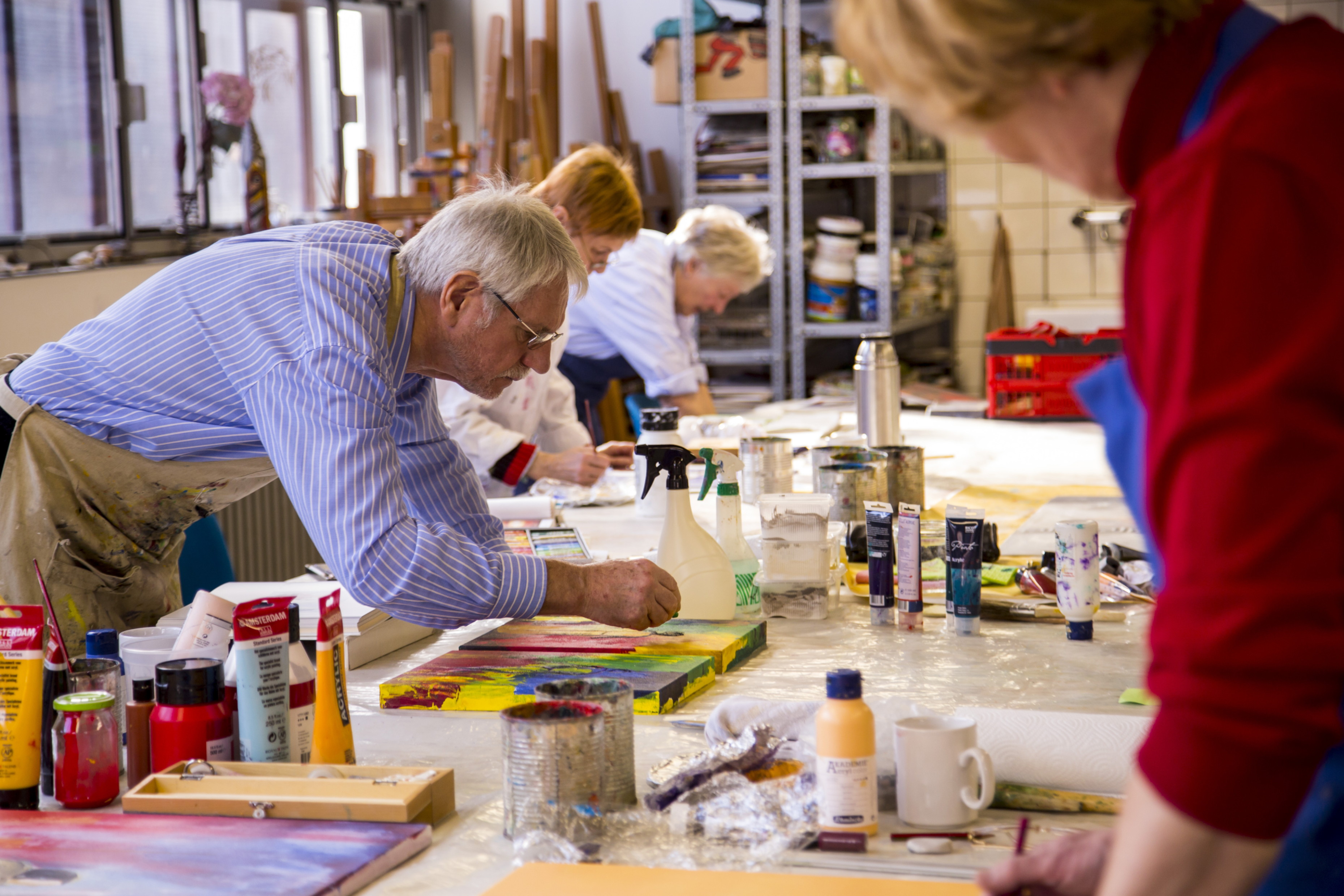Mehrere ältere Personen arbeiten konzentriert an einem langen Tisch in einem hellen, gut ausgestatteten Kunstatelier. Sie malen auf Leinwänden mit bunten Acrylfarben. Im Vordergrund steht ein Mann mit grauem Haar und Brille, der sich über sein Werk beugt. Auf dem Tisch liegen zahlreiche Malutensilien: Pinsel, Farbtuben, Becher, Lappen und eine Sprühflasche. Regale mit weiteren Materialien stehen im Hintergrund. Die Stimmung wirkt kreativ und gemeinschaftlich.