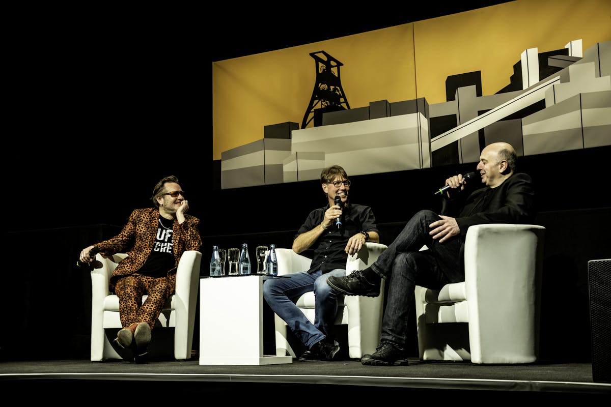 Auf einer Bühne sitzen drei Männer in weißen Sesseln und führen ein lebendiges Gespräch im Rahmen des Talkformats #halbzwölf auf dem Welterbe Zollverein in Essen. In der Mitte moderiert Peter Großmann das Gespräch. Rechts sitzt der bekannte Kabarettist und Autor Frank Goosen, leger in schwarzer Kleidung, mit Mikrofon in der Hand und einem Lächeln im Gesicht. Links sitzt ein Mann in einem auffälligen, orange-schwarz gemusterten Anzug, entspannt mit dem Kopf aufgestützt. Hinter ihnen hängt ein großformatiges, stilisiertes Bild der Zeche Zollverein mit dem ikonischen Doppelbock-Förderturm auf orangem Hintergrund.