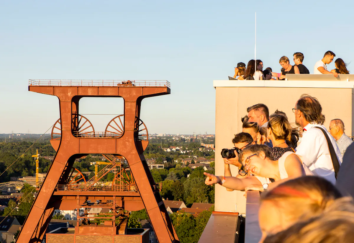Eine Gruppe von Besucherinnen und Besuchern steht auf der Dachterrasse des Ruhr Museums auf dem UNESCO-Welterbe Zollverein in Essen. Sie blicken neugierig und begeistert in Richtung des markanten Doppelbocks – das ikonische rote Fördergerüst der ehemaligen Zeche. Die Szene spielt sich bei warmem Abendlicht ab, das die Gesichter der Menschen und das rostrote Industriedenkmal in goldenem Glanz erstrahlen lässt. Im Hintergrund ist die grüne Stadtlandschaft des Ruhrgebiets zu sehen. Eine Frau im Vordergrund zeigt lächelnd auf etwas am Horizont – ein Moment der geteilten Faszination für Industriekultur und Geschichte.