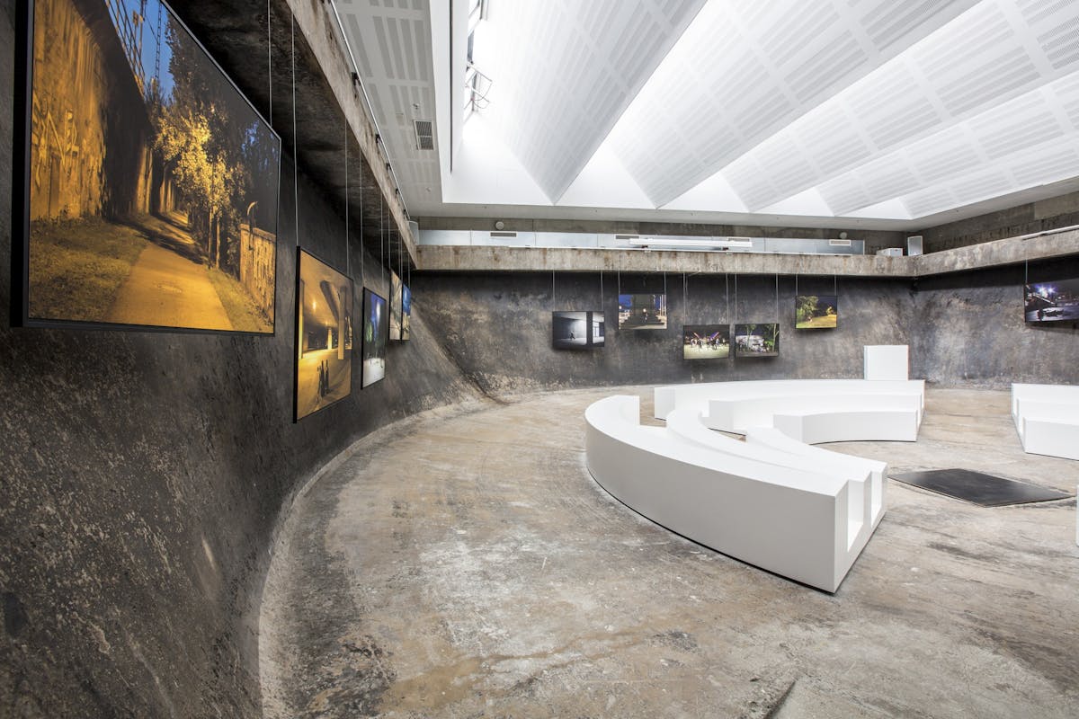 Innenaufnahme eines modernen Ausstellungsraums mit rauem, dunklem Betonboden und -wänden. Der Raum ist abgerundet und leicht abgesenkt wie ein Amphitheater. An den Wänden hängen großformatige Fotografien, die nächtliche Szenen im urbanen Raum zeigen, z. B. Straßenlaternen, Brückenunterführungen und menschenleere Wege. Die Decke ist hell und geometrisch strukturiert, mit indirekter Beleuchtung. In der Mitte des Raumes stehen weiße, gebogene Sitzbänke, die sich dem Verlauf der Wand anpassen und ein konzentrisches Halbrund bilden. Die Atmosphäre wirkt zugleich künstlerisch und industriell.