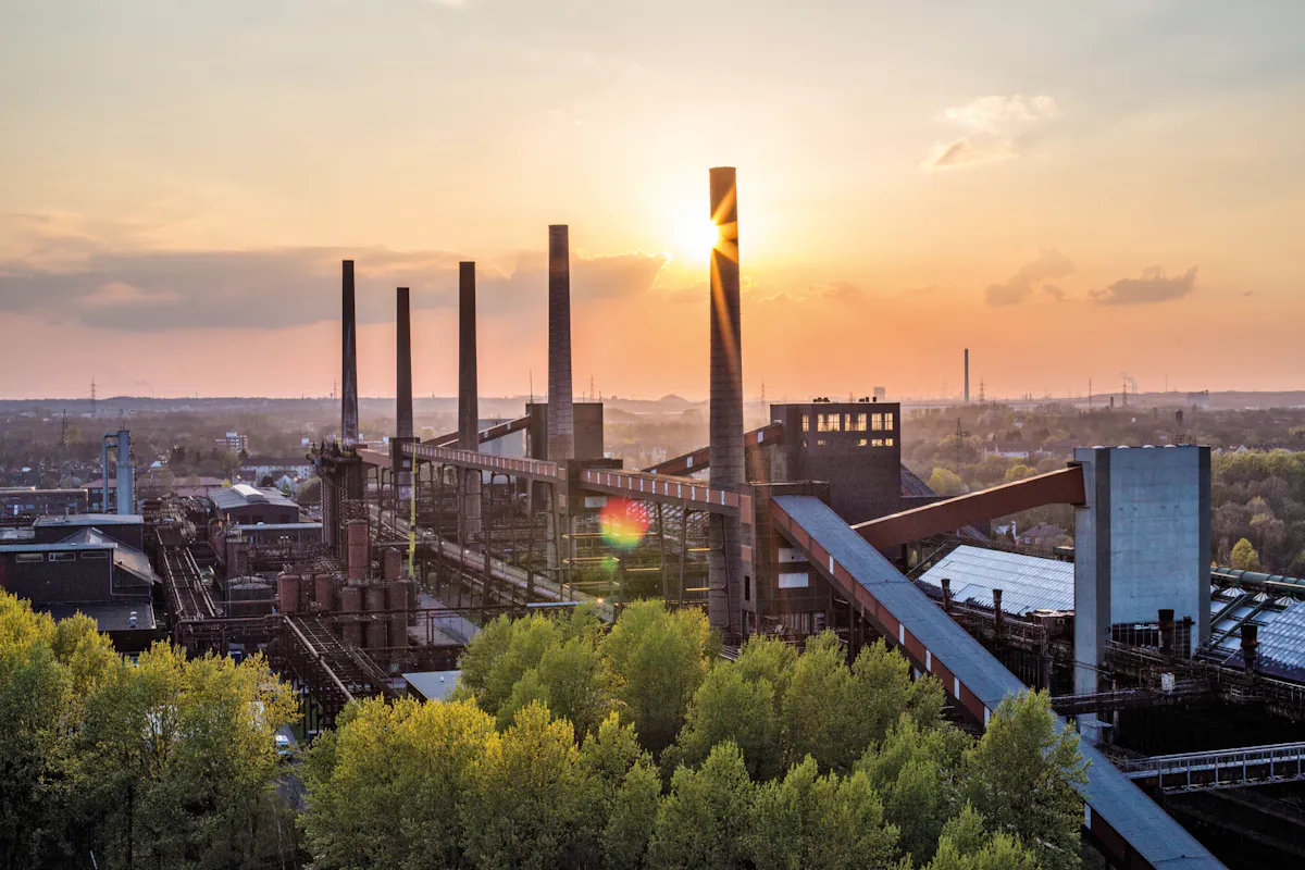 Panoramaaufnahme der Kokerei Zollverein bei Sonnenuntergang. Die industrielle Anlage mit ihren markanten, hohen Schornsteinen und rostrot-schwarzen Förderbändern erstreckt sich über das Bild. Die Sonne steht tief am Himmel und scheint direkt hinter einem Schornstein hervor, wodurch ein warmer Lichtschein entsteht. Im Vordergrund leuchten frische grüne Baumkronen, im Hintergrund erstreckt sich das flache Ruhrgebiet mit weiteren Industrieanlagen bis zum Horizont. Die Szene vermittelt einen ruhigen, fast poetischen Kontrast zwischen Industriearchitektur und Natur.