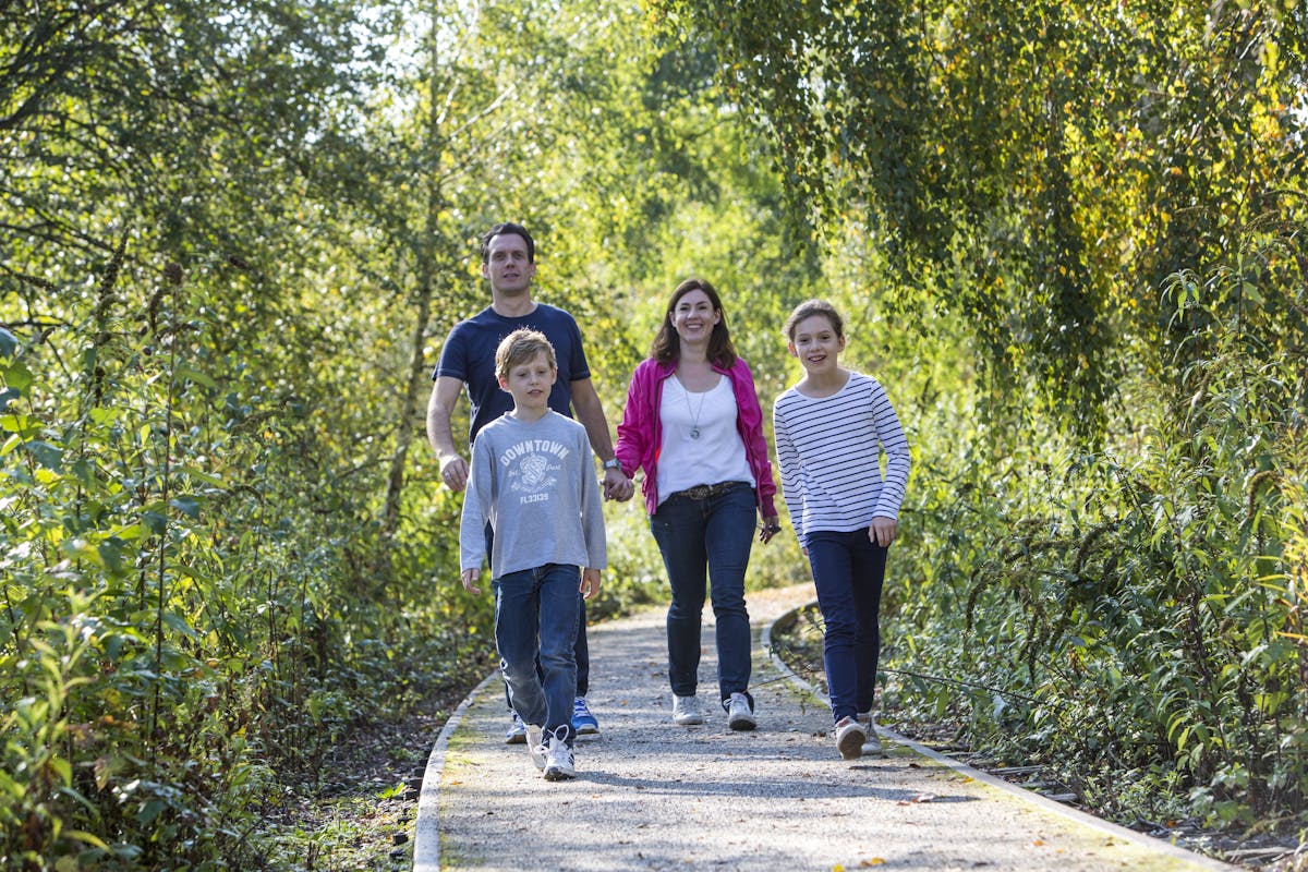 Eine vierköpfige Familie spaziert auf einem schmalen, geschwungenen Weg durch eine grüne, dicht bewachsene Naturlandschaft. Vorne gehen ein Junge mit grauem Pullover und eine lächelnde Jugendliche im weiß-blau gestreiften Shirt. Dahinter folgen ihre Eltern: der Vater trägt ein dunkelblaues T-Shirt, die Mutter ein weißes Shirt mit pinker Jacke. Die Sonne scheint durch das Laub, wodurch die Szene freundlich und lebendig wirkt. Der Weg ist mit feinem Kies bedeckt und wird von üppigem Grün gesäumt.