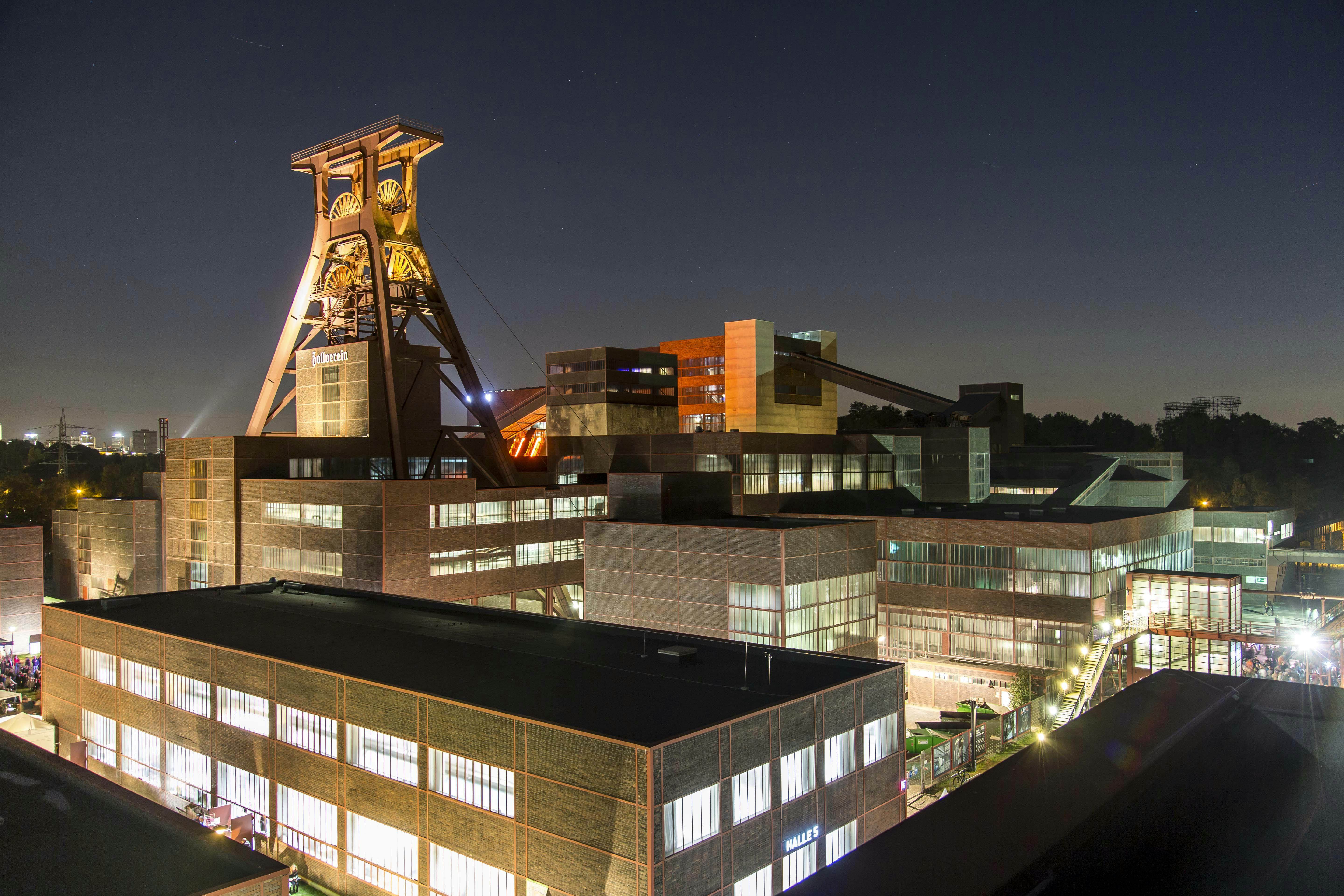 Nächtliche Panoramaaufnahme des UNESCO-Welterbes Zollverein in Essen. Im Zentrum steht das markante, angestrahlte Doppelbock-Fördergerüst des Schachts XII, auf dem der Schriftzug: Zollverein zu lesen ist. Die umliegenden Backsteinbauten der ehemaligen Zeche sind großflächig erleuchtet, viele Fenster strahlen hell in die Dunkelheit. Im Vordergrund ist die Halle 5 zu erkennen, im Hintergrund leuchtet das orangefarbene Rolltreppenhaus. Der Himmel ist klar und dunkel, mit wenigen Sternen sichtbar. Die Szene vermittelt industrielle Monumentalität und moderne kulturelle Nutzung bei Nacht.