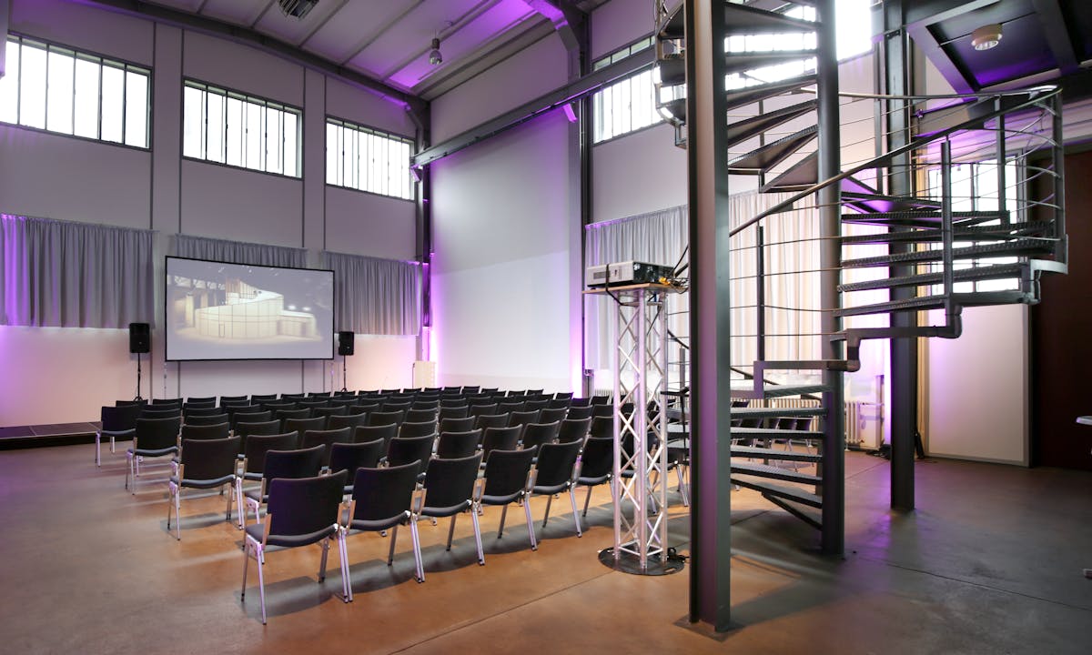Innenansicht eines modernen Veranstaltungsraums mit hoher Decke und industriellem Flair. In der Bildmitte befinden sich mehrere Reihen schwarzer Stühle, die auf eine große Projektionsleinwand ausgerichtet sind. Auf der Leinwand ist ein Standbild einer Architektur-Visualisierung zu sehen. Der Raum wird von lila Lichtakzenten beleuchtet, die an den Wänden und Deckenstrukturen sichtbar sind. Rechts im Bild führt eine schwarze Wendeltreppe aus Metall in ein Obergeschoss. Im Vordergrund steht ein silbernes Traversenstativ mit einem Beamer. Der Raum ist hell, offen und für Vorträge oder Konferenzen eingerichtet.