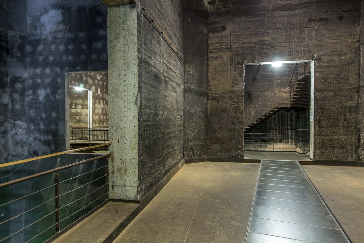 Blick in das Innere eines ehemaligen Industriegebäudes mit hohen, dunklen Betonwänden. Im Zentrum verläuft ein begehbarer Steg oder eine Rampe mit Metallplatten, der durch breite Durchgänge in angrenzende Räume führt. In einem dieser Räume ist eine Treppe mit Metallgeländer sichtbar, die schräg nach oben führt. Die gesamte Szenerie wirkt roh, industriell und ursprünglich belassen – typische Architektur der Zeche Zollverein. Links und rechts sind Geländer montiert, um Absturz zu verhindern. Die Lichtstimmung ist gedämpft, aber durch künstliche Deckenleuchten erhellt.