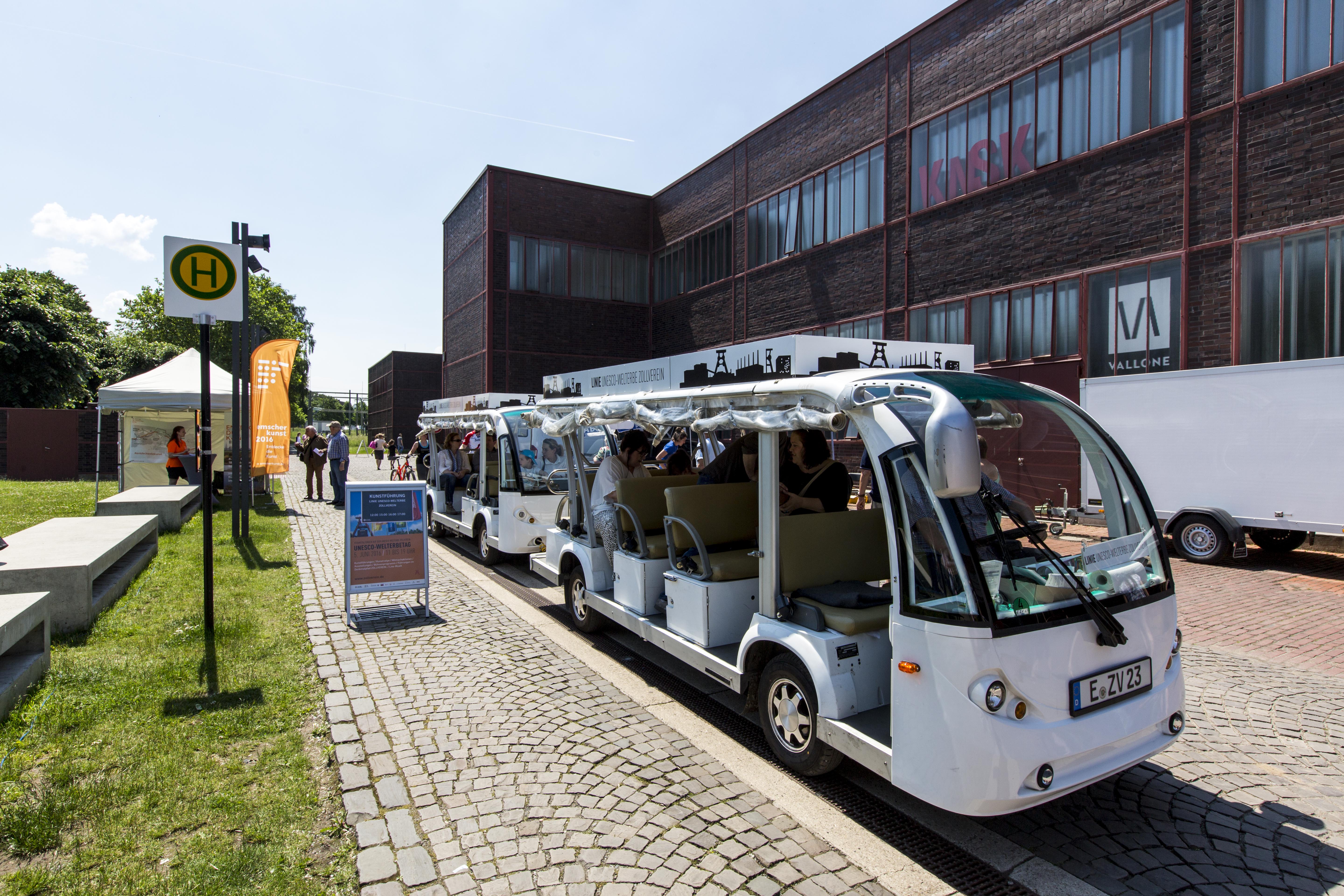 Zwei weiße Besucher-Shuttles stehen an einer Haltestelle auf dem Gelände des Welterbes Zollverein. Im Hintergrund ist Halle 6 mit roter Backsteinfassade und großen Fenstern zu sehen. Menschen steigen ein.