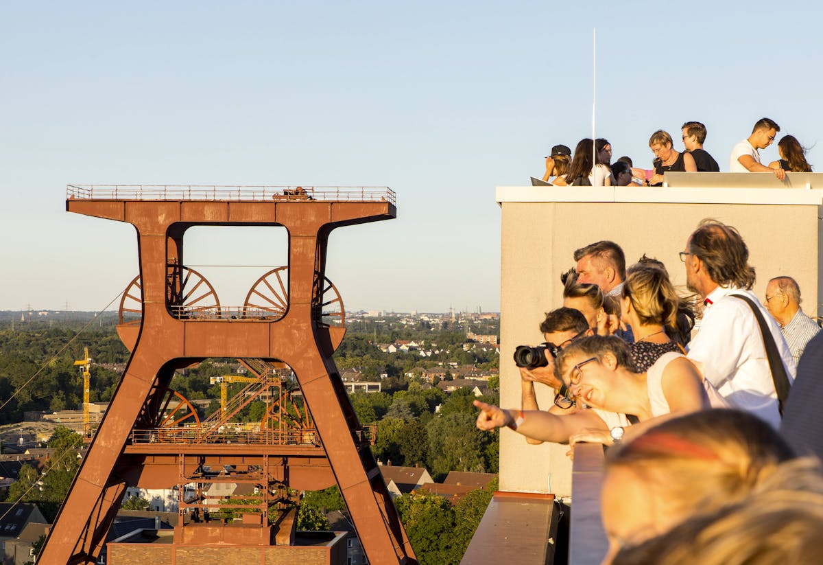 Eine Gruppe von Besucherinnen und Besuchern steht auf der Dachterrasse des Ruhr Museums auf dem UNESCO-Welterbe Zollverein in Essen. Sie blicken neugierig und begeistert in Richtung des markanten Doppelbocks – das ikonische rote Fördergerüst der ehemaligen Zeche. Die Szene spielt sich bei warmem Abendlicht ab, das die Gesichter der Menschen und das rostrote Industriedenkmal in goldenem Glanz erstrahlen lässt. Im Hintergrund ist die grüne Stadtlandschaft des Ruhrgebiets zu sehen. Eine Frau im Vordergrund zeigt lächelnd auf etwas am Horizont – ein Moment der geteilten Faszination für Industriekultur und Geschichte.