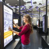 Eine junge Frau mit langen, offenen Haaren und rotem Pullover interagiert mit einem großen Touchscreen im Portal der Industriekultur auf dem Gelände der Zeche Zollverein. Auf dem Bildschirm ist eine historische Schwarzweißaufnahme der Kokerei Hansa in Dortmund zu sehen, darüber der Text: Kokerei Hansa Dortmund, darunter in einem gelben Balken: Geschichte 1920. Im Hintergrund sind weitere interaktive Stationen mit Bildschirmen zu sehen. Die Decke ist mit stilisierten Kartenfragmenten und blauen Lichtlinien gestaltet. Die Atmosphäre ist modern, interaktiv und bildungsorientiert.