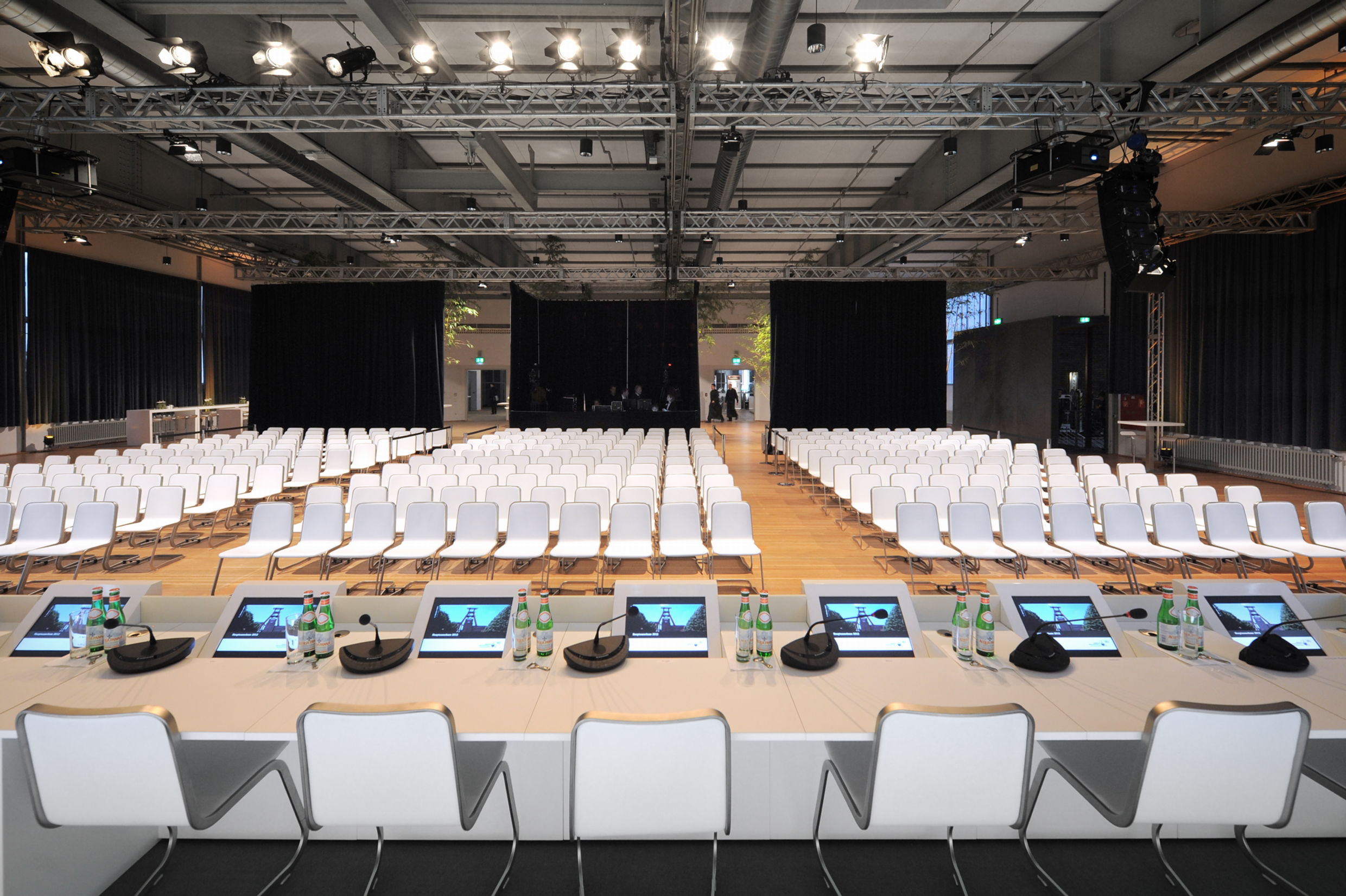 Großer, moderner Konferenzsaal mit Reihen weißer Stühle, die auf eine Bühne am anderen Ende des Raums ausgerichtet sind. Im Vordergrund befindet sich ein langes Podium mit weißen Stühlen für die Redner:innen. Auf dem Tisch stehen mehrere Mikrofone, Flaschen mit Mineralwasser und Monitore, die das Logo oder ein Bild von Zeche Zollverein zeigen. Der Raum ist technisch voll ausgestattet mit Licht- und Tontechnik, sichtbar an Traversen unter der Decke. Schwarze Vorhänge und dezente Beleuchtung geben dem Raum eine professionelle und elegante Atmosphäre.