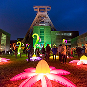 Nächtliche Lichtinstallation auf dem Gelände der Zeche Zollverein vor dem beleuchteten Doppelbock-Fördergerüst. Auf der Wiese vor dem Gebäude sind leuchtende, übergroße Blumen und fantasievolle Pflanzen in kräftigen Farben wie Pink, Gelb und Grün installiert. Zahlreiche Menschen spazieren zwischen den Lichtobjekten umher und bestaunen die Szenerie. Die Fassade des Förderturms ist in grünem Licht angestrahlt, die Atmosphäre wirkt lebendig und märchenhaft.